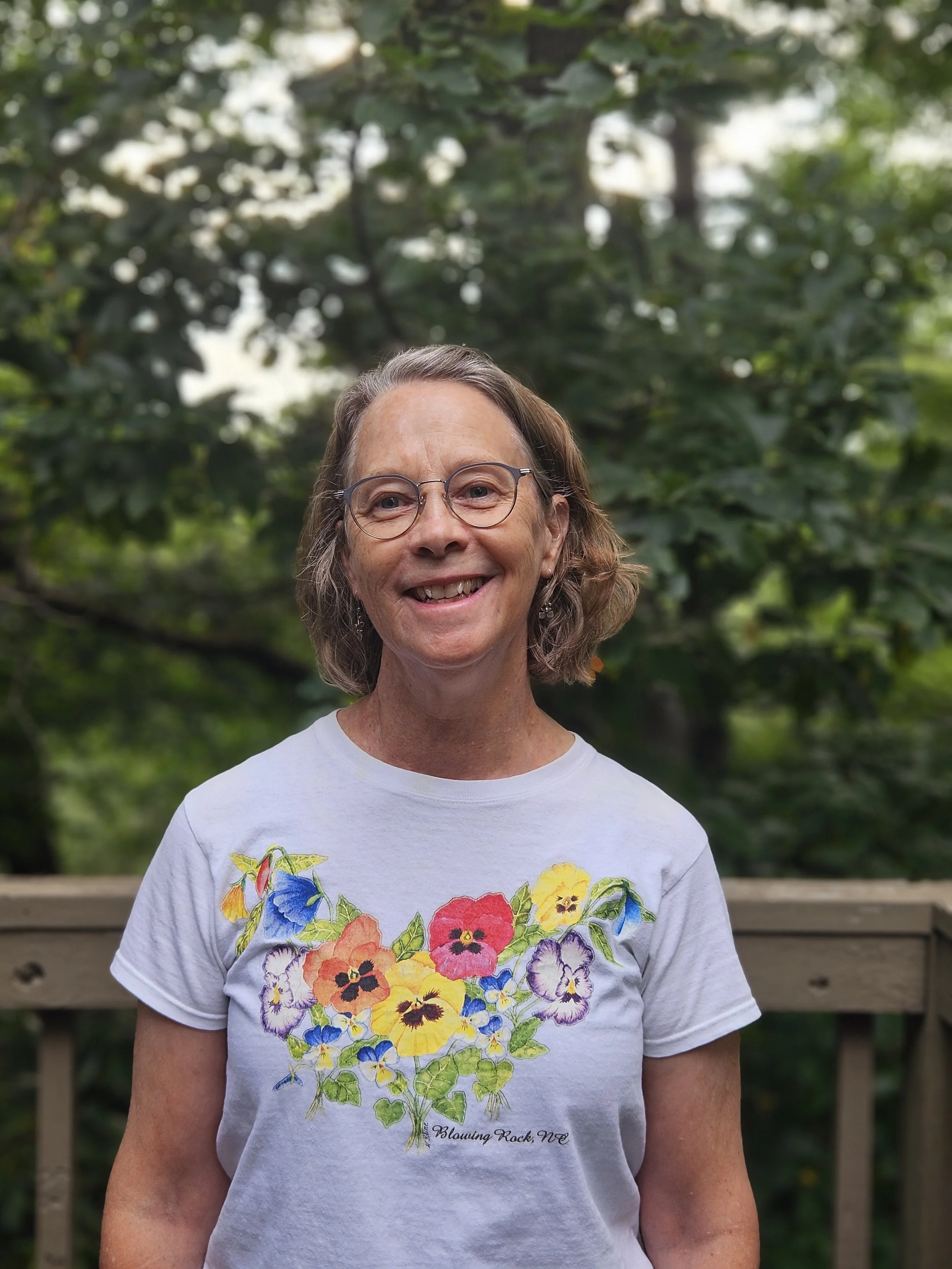 A woman with glasses smiling outdoors in front of a green, leafy background. She is wearing a white T-shirt with colorful flowers and the text "Blowing Rock, NC" on it.