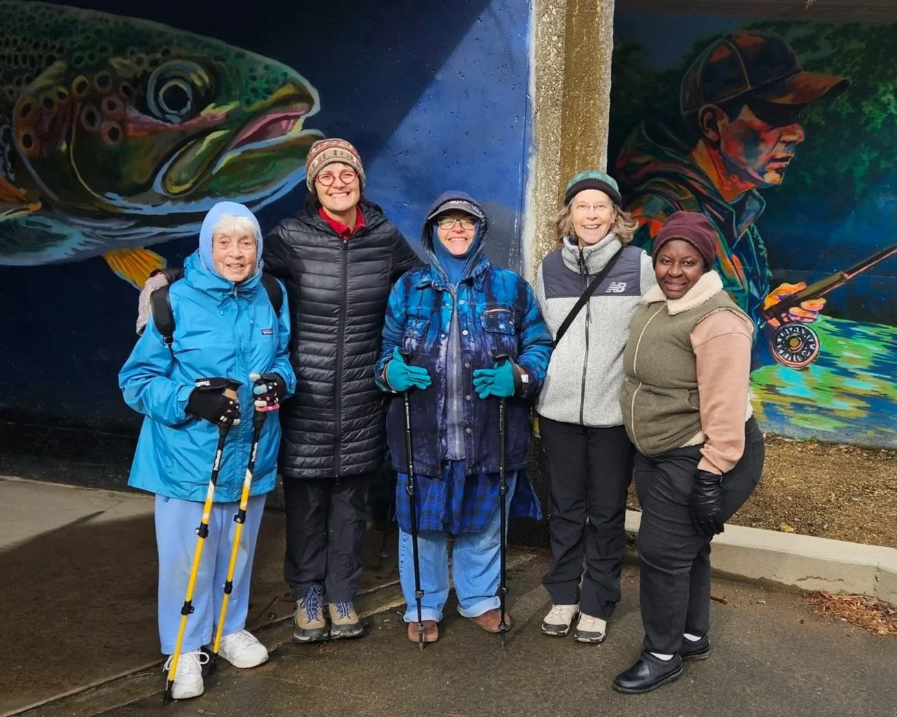 Group of five women dressed in outdoor winter gear, standing in front of colorful mural paintings of a fish and a fisherman, smiling and holding walking sticks.