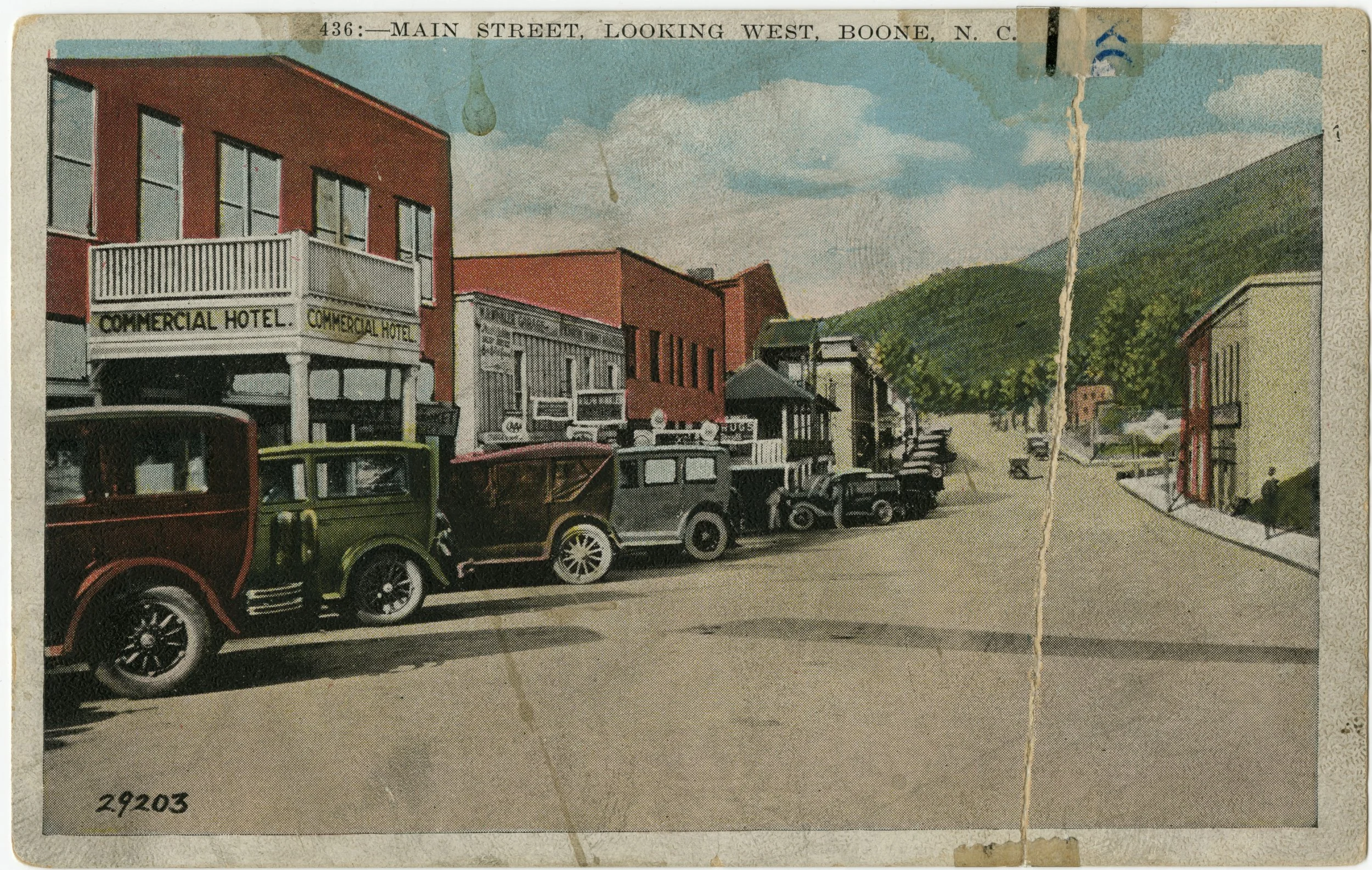 Vintage postcard of Main Street, looking west in Boone, North Carolina. Features early 20th-century cars parked along the street, with a red building labeled Commercial Hotel and additional buildings with signs and balconies. Mountains are visible in the background under a partly cloudy sky.