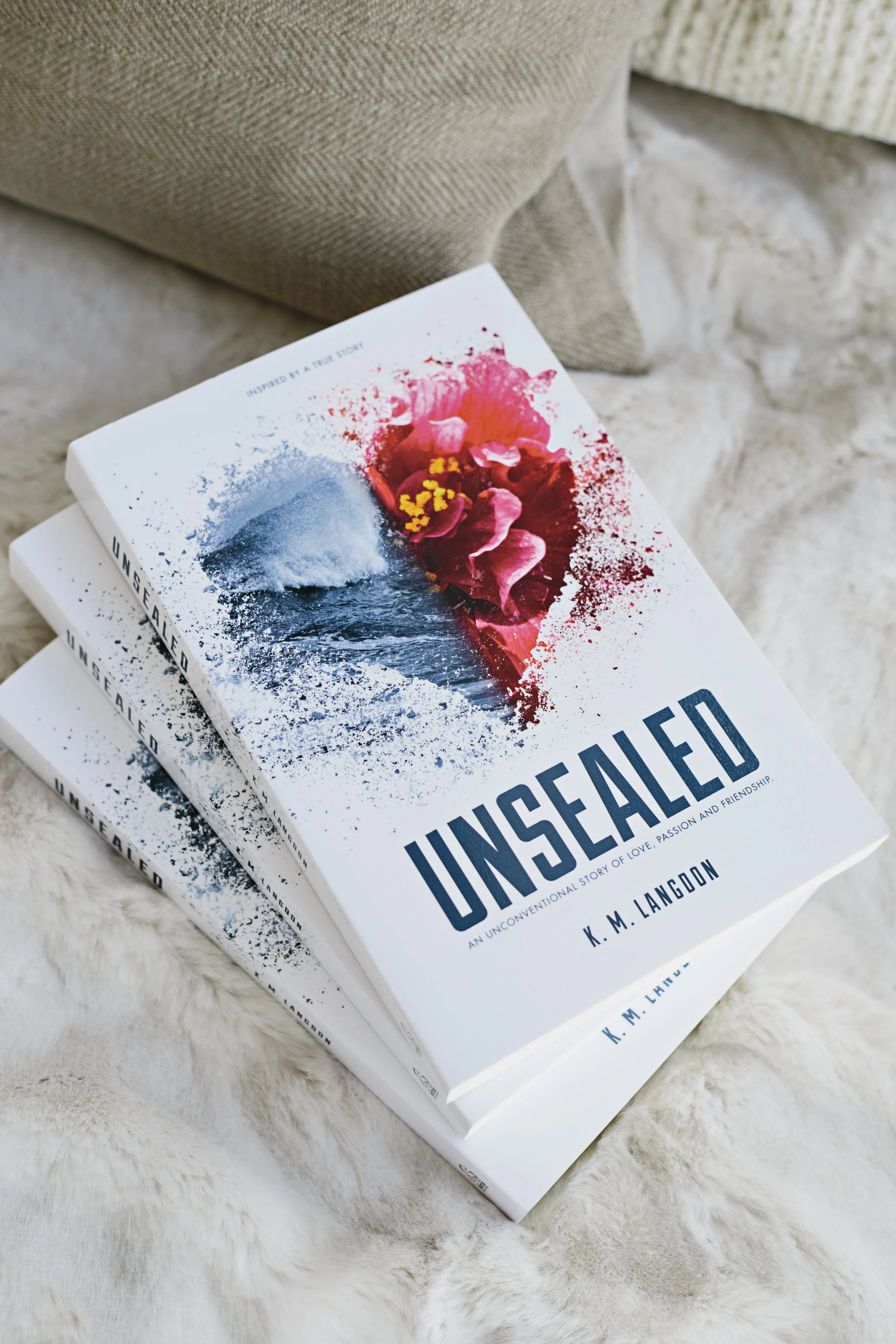 Two books titled 'Unsealed' with a cover design featuring a sailboat, a large pink flower, and splashes of color, resting on a fluffy, beige surface.