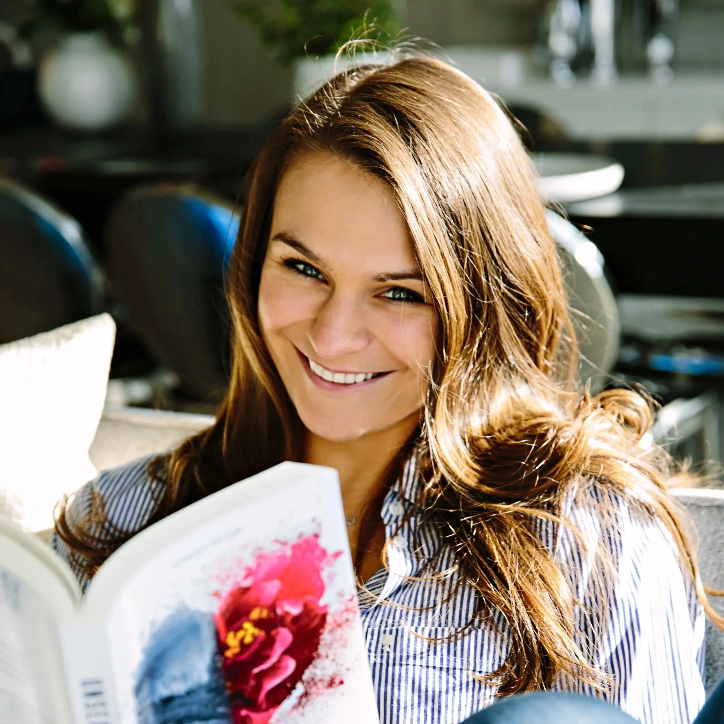 A woman with long brown hair smiling, wearing a stripe shirt, holding a bouquet of pink and red flowers.