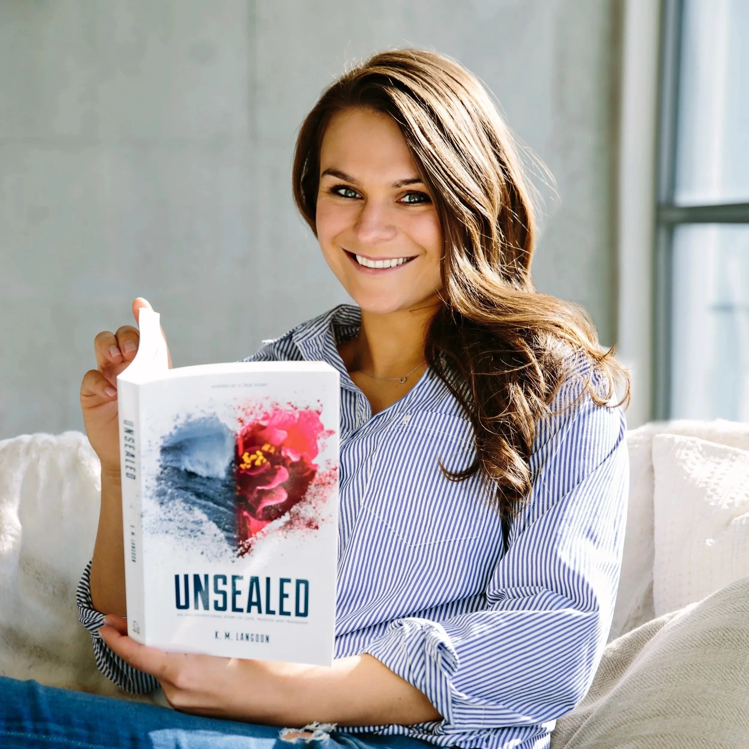 A woman with long brown hair and blue eyes smiling while holding a book titled 'Unsealed' in front of her. She is sitting on a couch in a room with a window visible in the background.