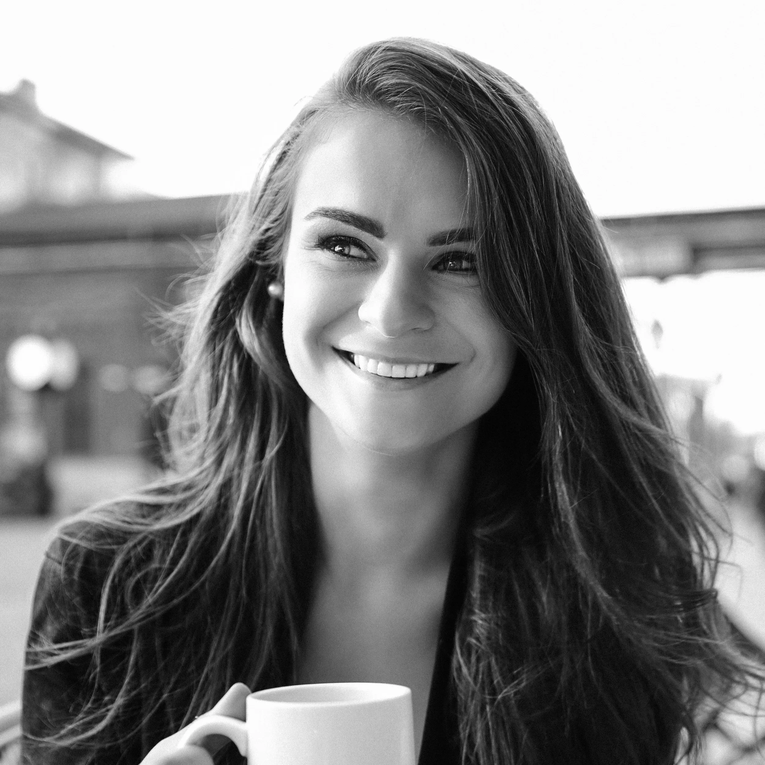 A black-and-white close-up photo of a woman's face. She is smiling, with her teeth showing, and holding a white mug near her mouth. Her hair is long, wavy, and cascades over her shoulder. The background appears to be outdoor with a blurred building or structure.