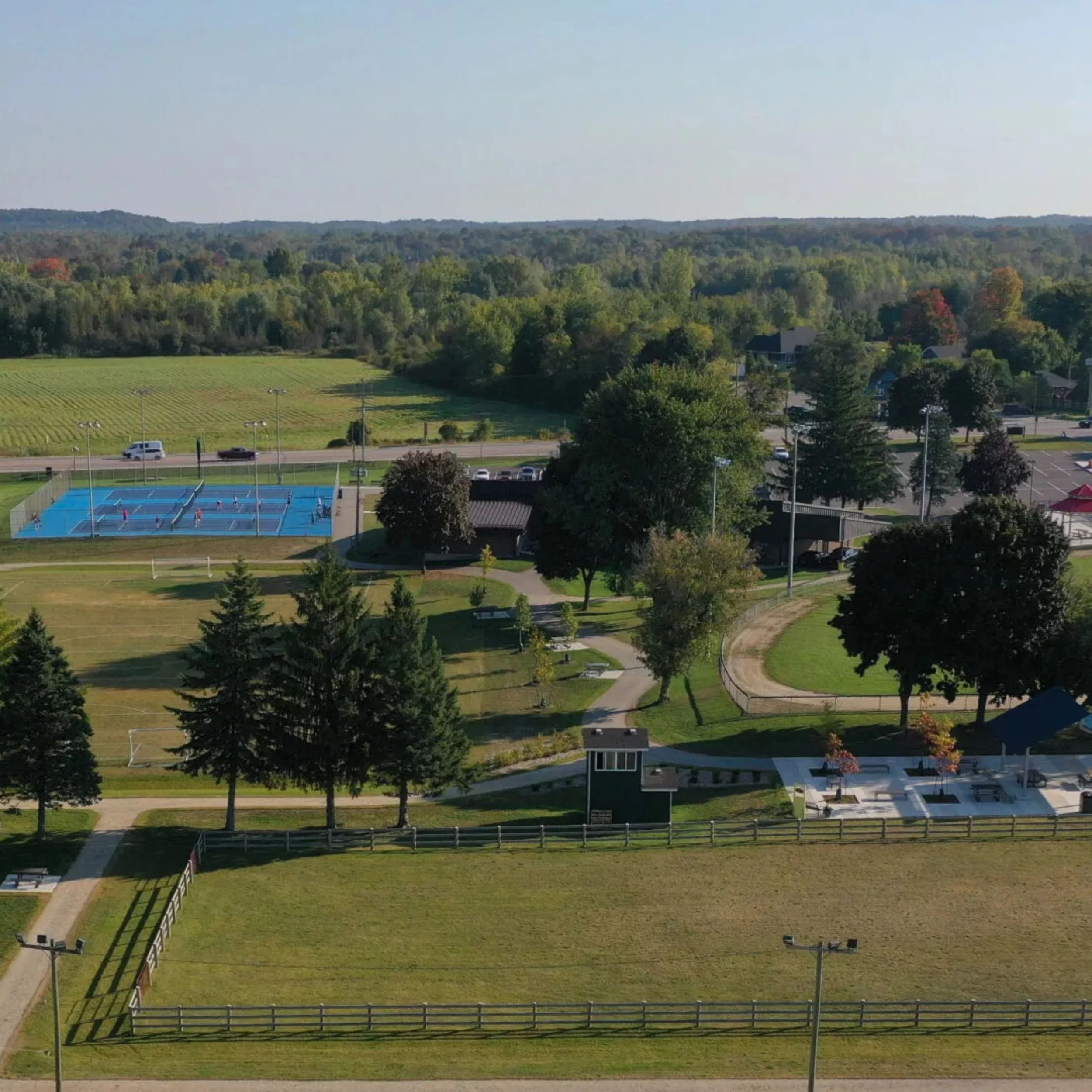 Aerial view of a park with tennis courts, grassy fields, trees, and a parking lot with cars, under a clear blue sky.