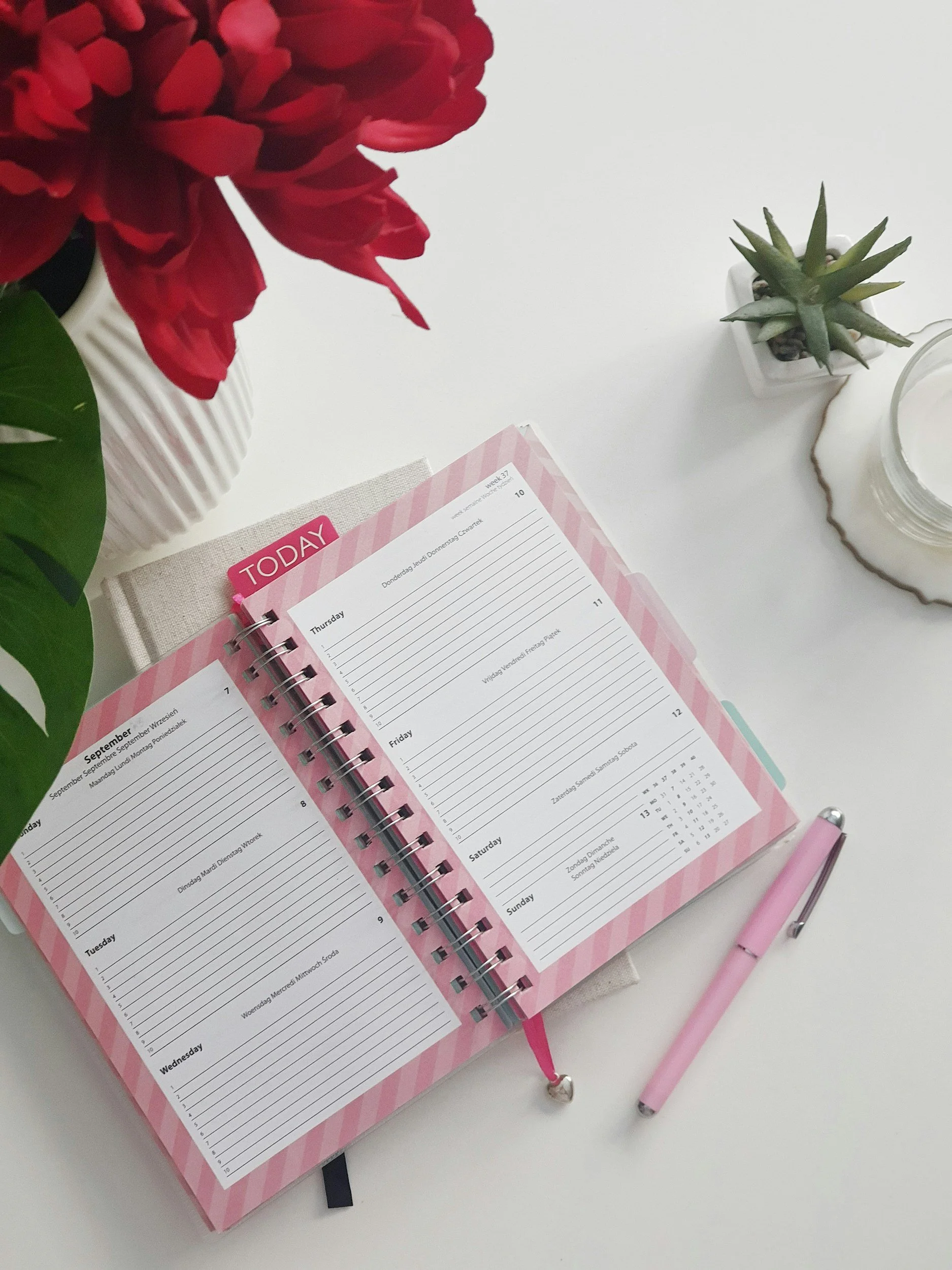 A planner open to the week of September 4-10, with days listed from Thursday to Wednesday. The planner has a pink striped border and white spaces for writing. A pink pen with a heart-shaped top is placed on the right side, and a white candle and a small potted succulent plant are also on the white surface. Part of a red flower in a white vase with green leaves is visible in the upper left corner.