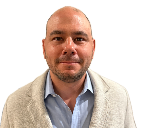 Headshot of a bald man with a beard, wearing a light blue collared shirt and a beige blazer, smiling against a white background.