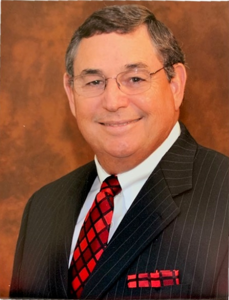 A smiling man in a dark pinstripe suit, white shirt, and red patterned tie, with glasses, against a brown background.