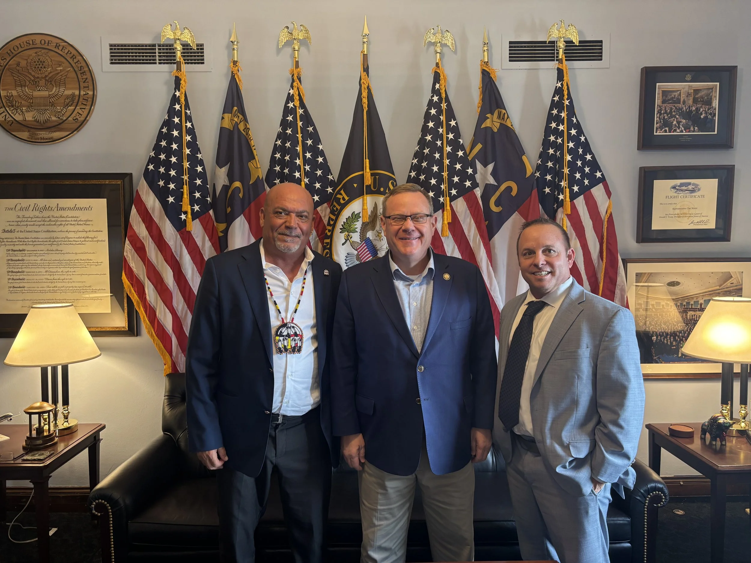 Three men in suits standing together in an office with multiple American flags and framed documents on the wall behind them.