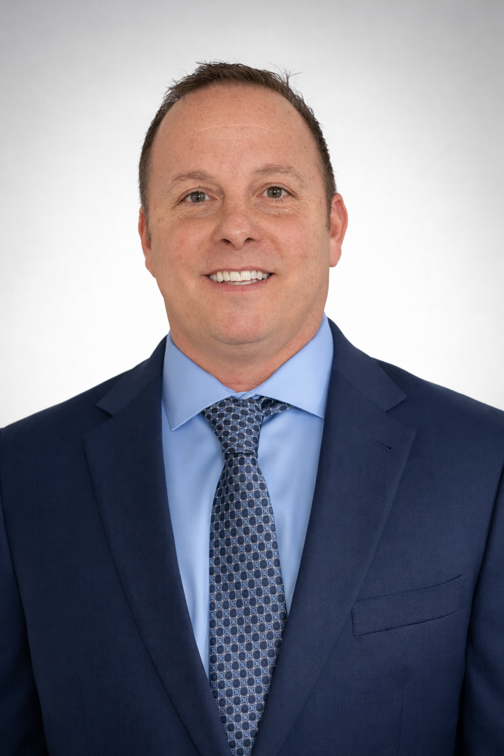 Headshot of a smiling man in a blue suit, light blue shirt, and patterned tie, against a plain background.