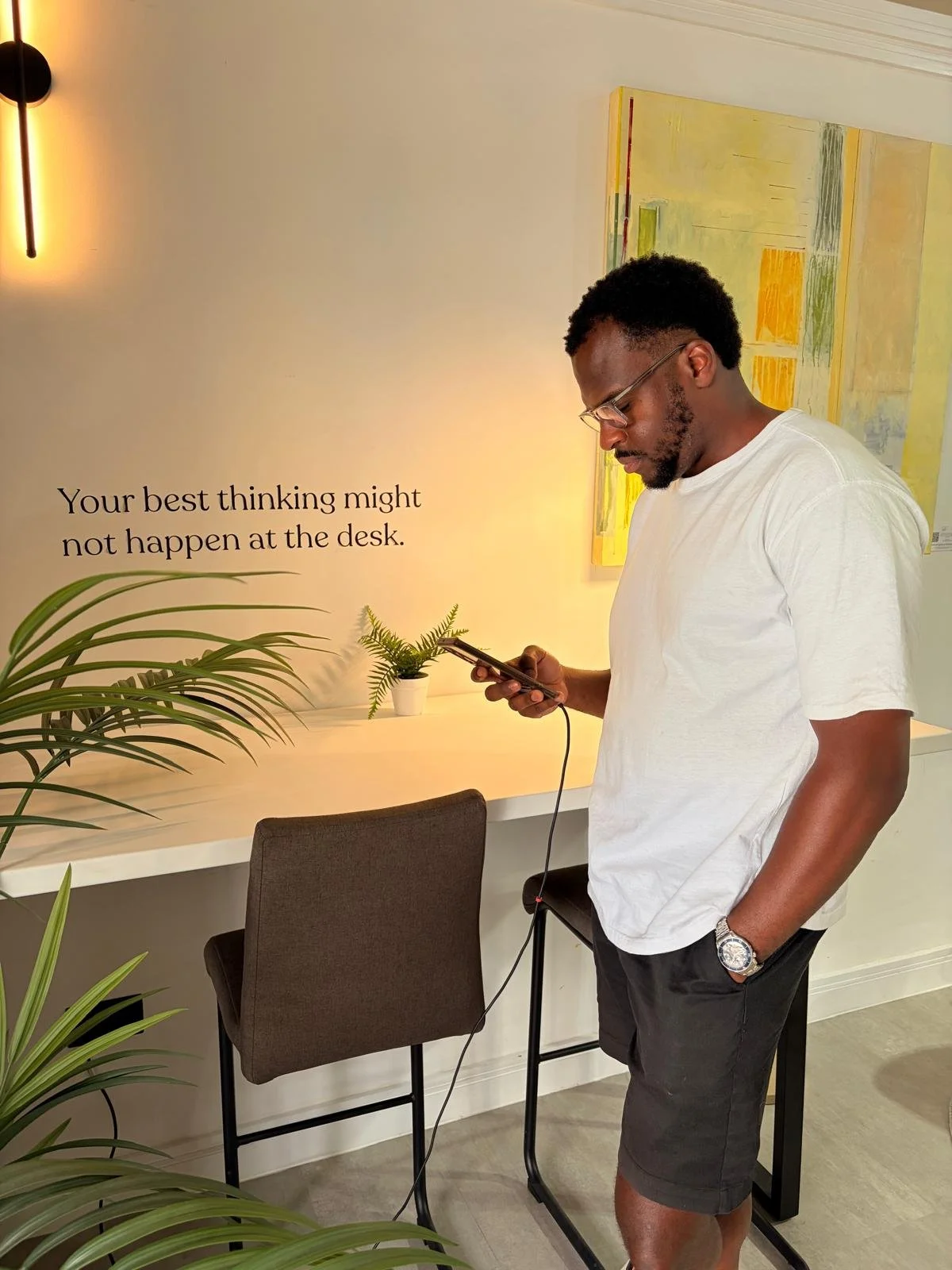 A man wearing a white T-shirt and glasses is standing next to a white counter, looking at his phone, with a wall decoration that includes a quote: "Your best thinking might not happen at the desk." There is a chair in front of the counter, some plants, and a yellow abstract painting on the wall.