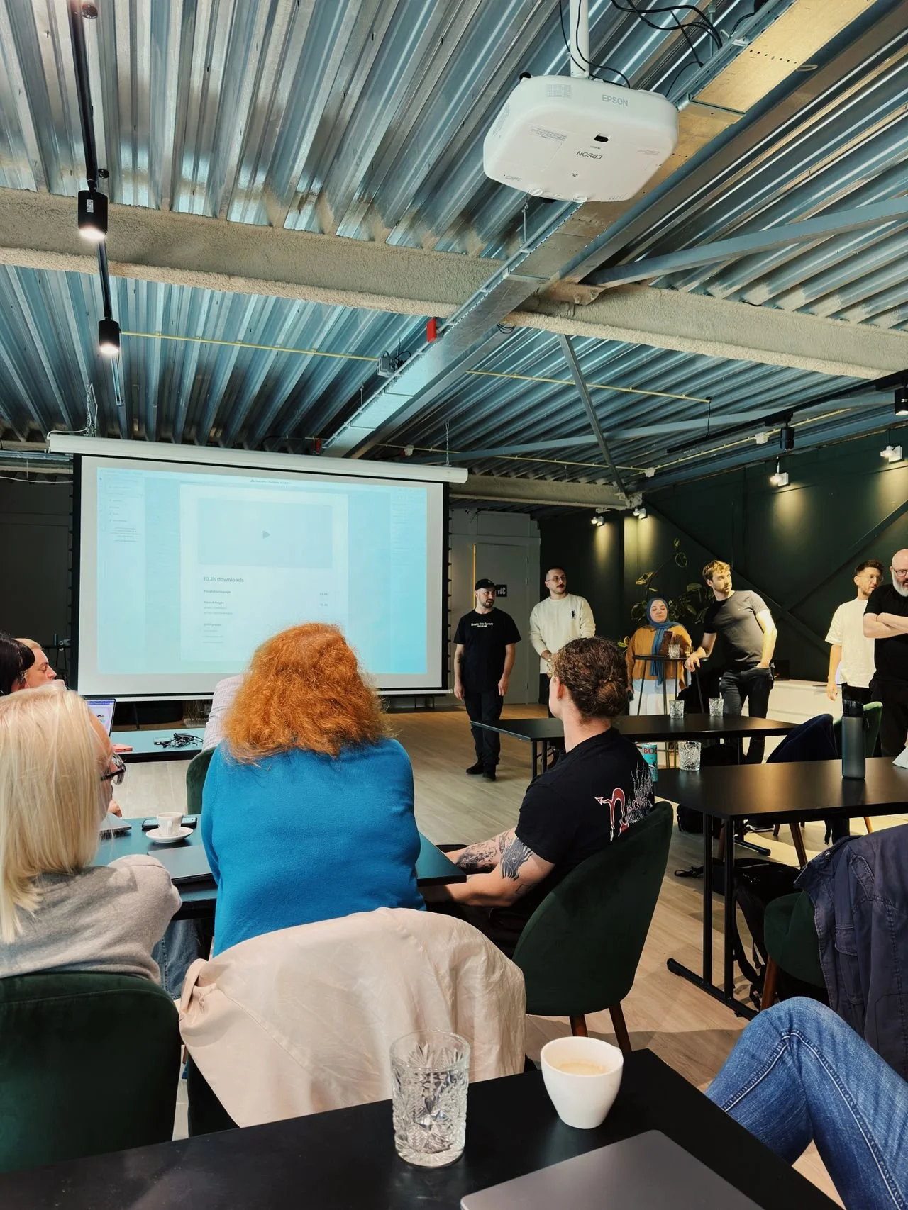 A group of people attending a presentation in a modern, industrial-style room with a metal ceiling and exposed pipes. Some are seated at tables with cups and glasses, while others stand near a large projection screen. Six individuals are standing at the front of the room, probably presenting or speaking, with a large screen displaying a webpage or app interface.