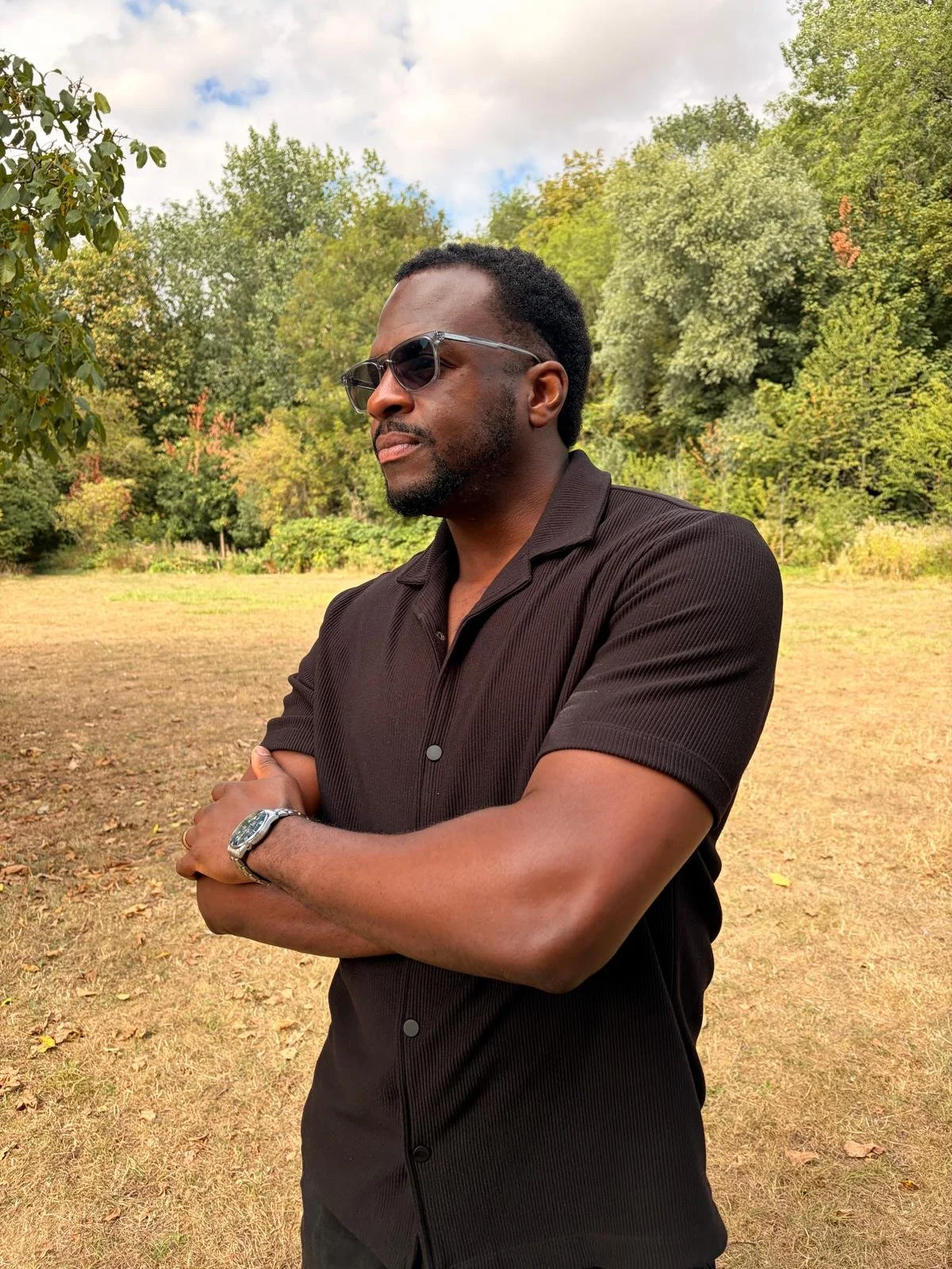 A man wearing black sunglasses and a dark short-sleeved button-up shirt, standing outdoors with crossed arms, in a park-like setting with trees and a partly cloudy sky.