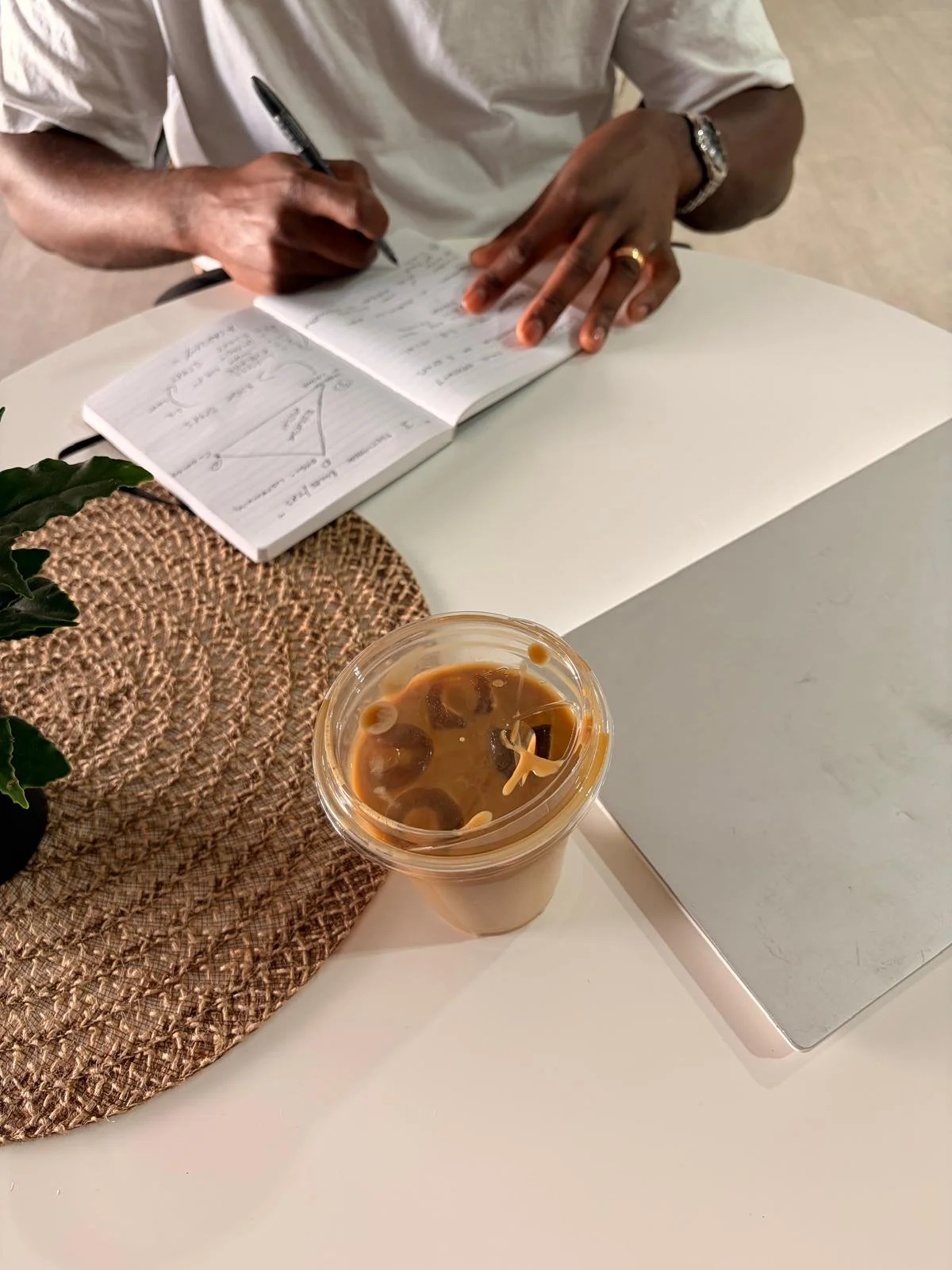 A person writing in a notebook at a white table, with a cup of iced coffee nearby, a plant on a woven placemat, and a laptop