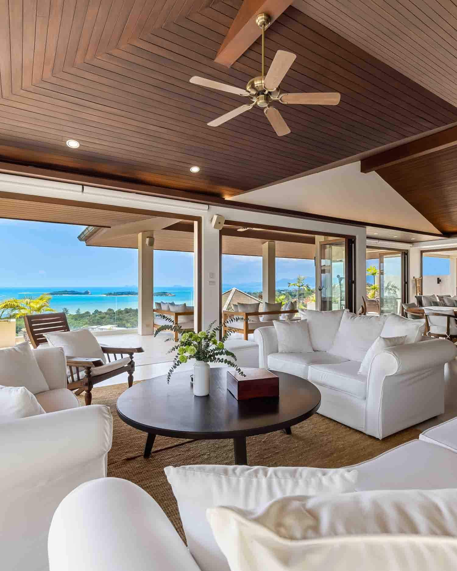 Spacious open-plan living room at Saensook Villa featuring plush white sofas, a dark wood coffee table, and sliding glass doors framing a panoramic ocean view.