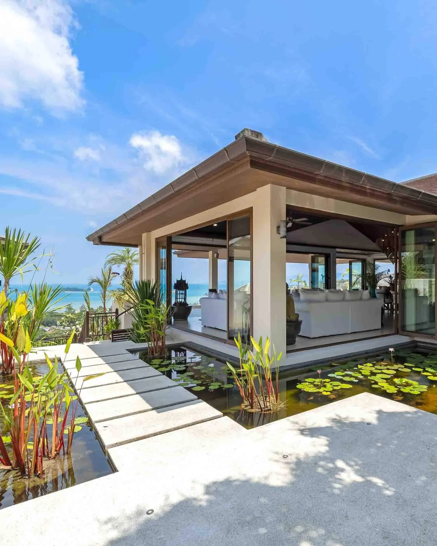 Modern entrance of Saensook Villa featuring stone stepping stones over a serene koi pond and water lilies with sea views beyond.