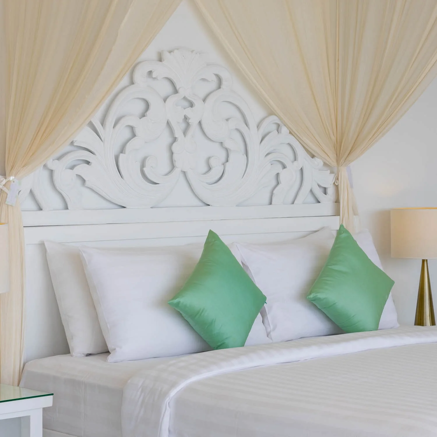Detailed shot of a white carved wooden headboard with elegant sheer draping and green accent pillows on a king bed at Saensook villa