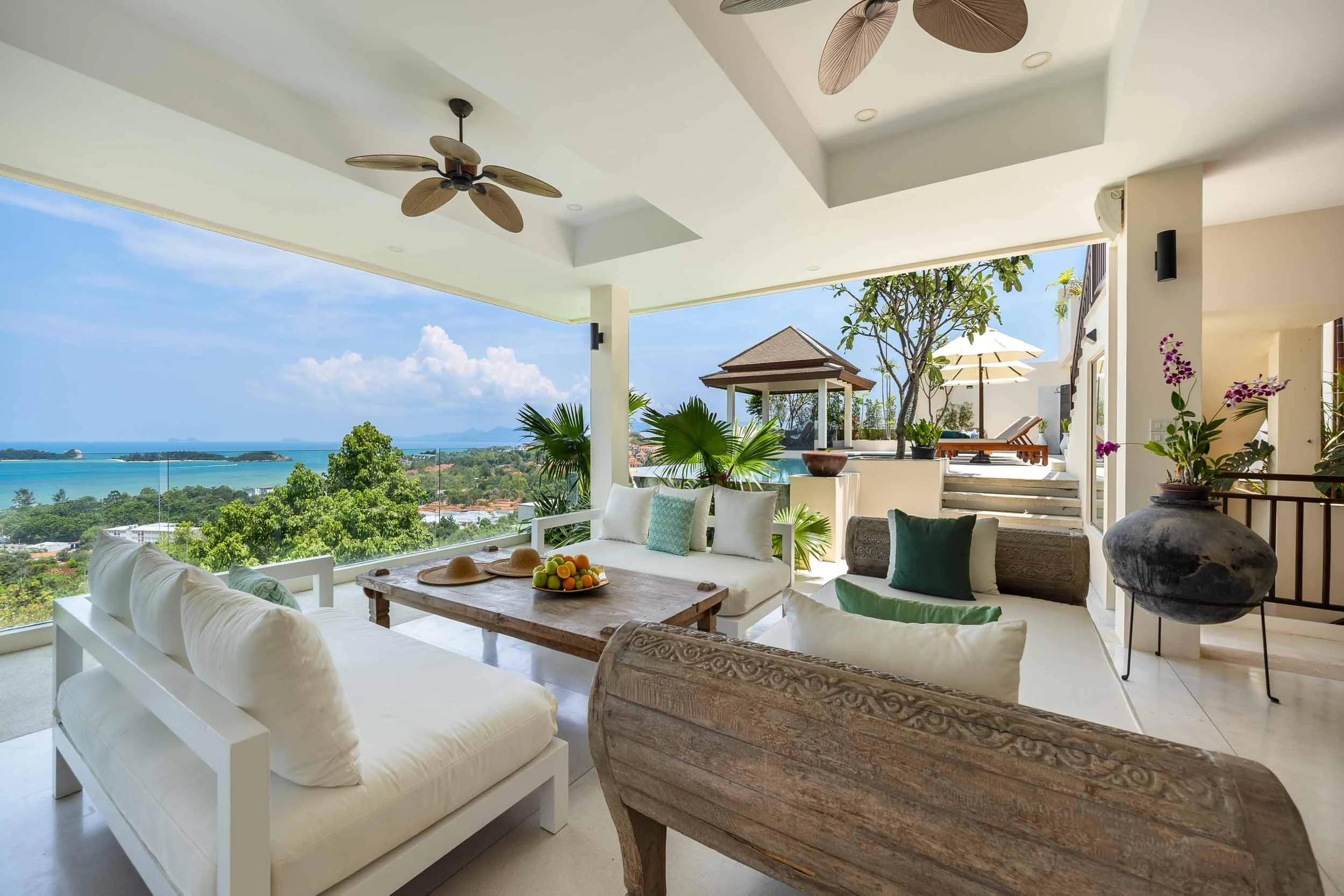 Open-air shaded terrace lounge at Saensook Villa with comfortable white couches, a rustic wooden coffee table, and ceiling fans overlooking the coastline.
