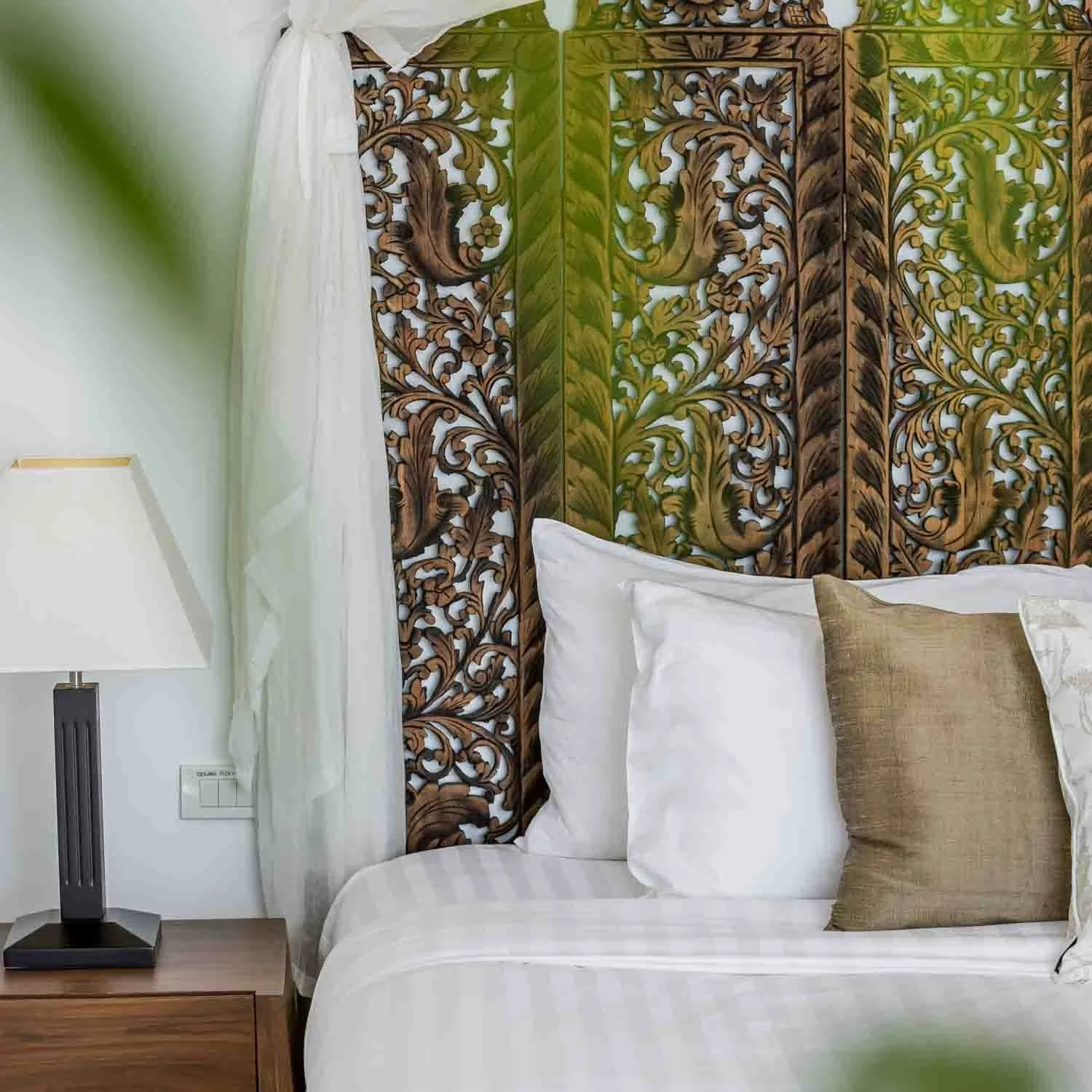 Close-up of an intricate traditional carved wood panel headboard behind crisp white linens in a bedroom suite at Saensook villa