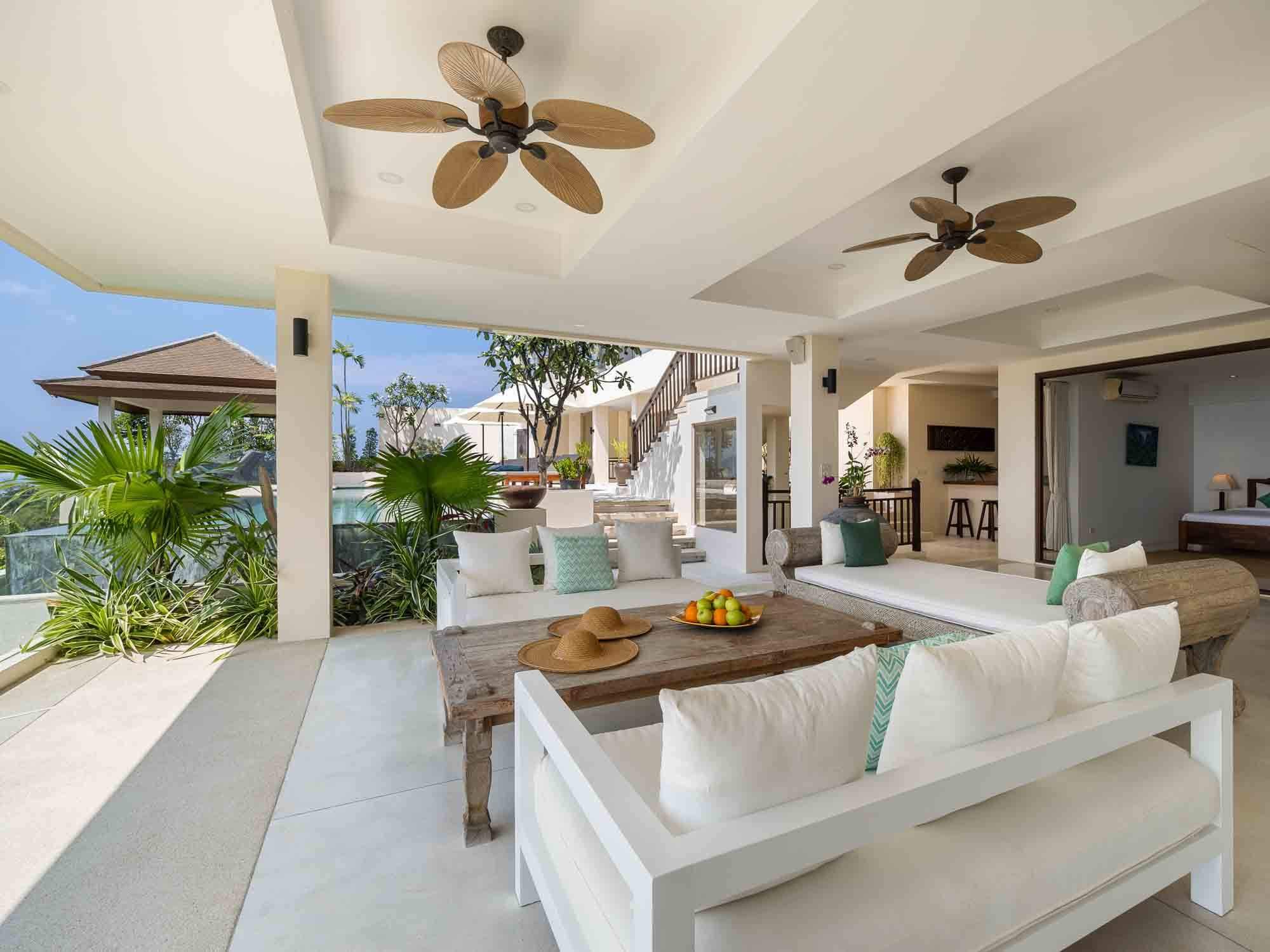 Covered outdoor lounge terrace furnished with white sofas, wooden coffee tables, and ceiling fans overlooking the pool and tropical greenery at Saensook villa