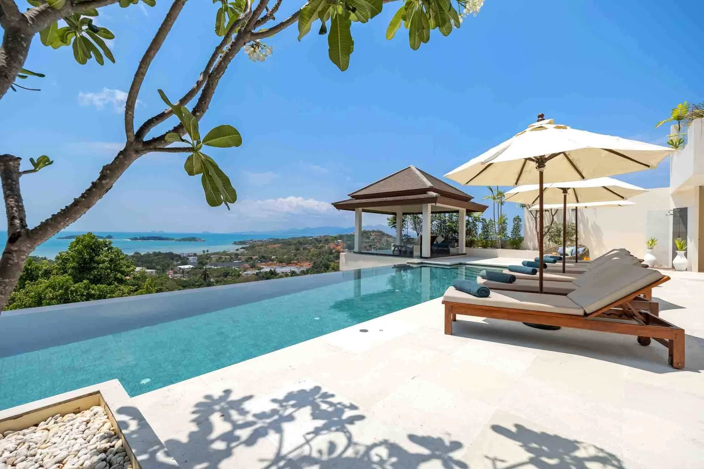 A luxurious outdoor pool at a modern villa with a scenic view of the ocean, lush greenery, and blue sky. There are lounge chairs with rolled towels, large white umbrellas, a shaded pavilion, and decorative plants.