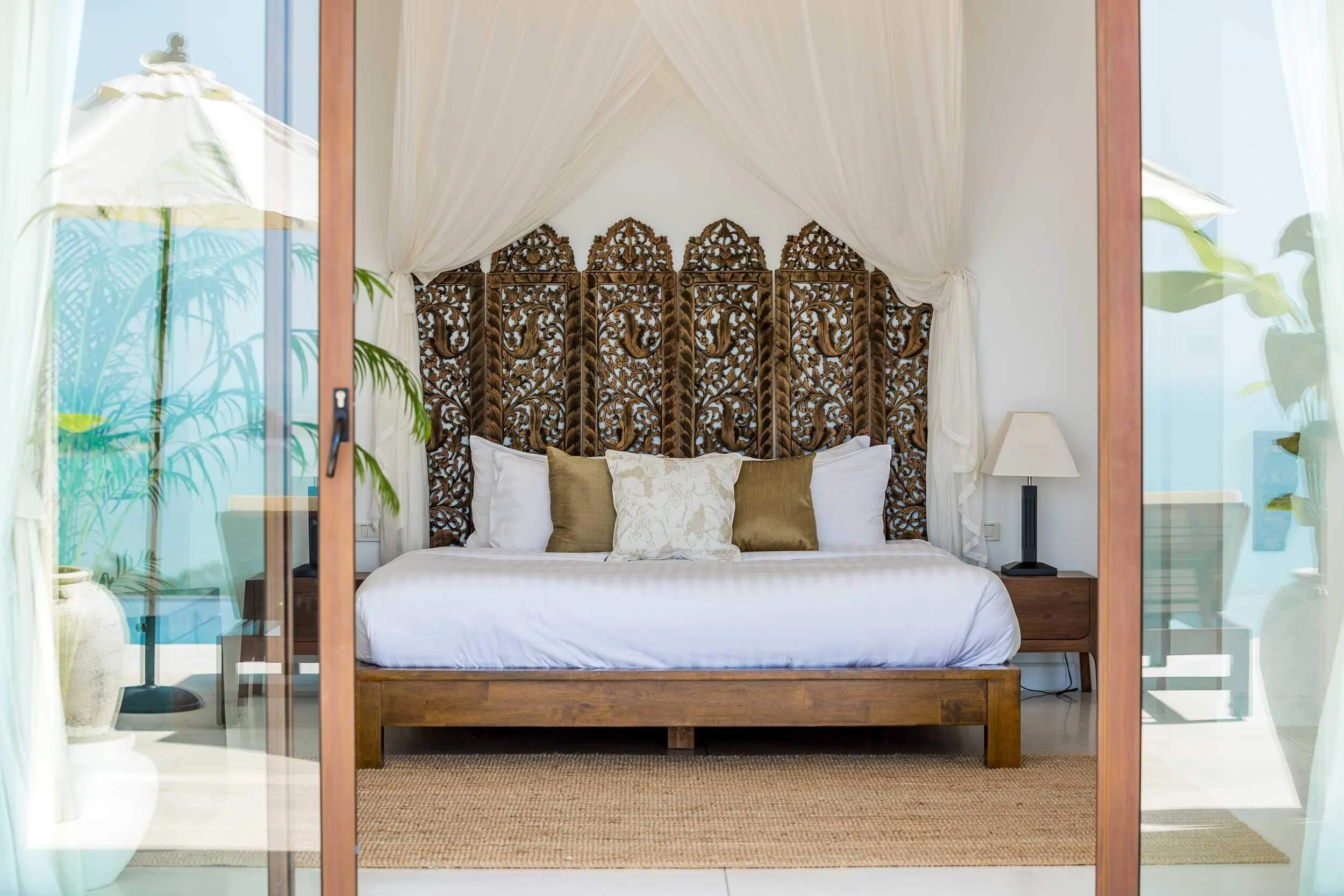 King-size bed featuring an ornate traditional carved dark wood headboard and white canopy framing pool and ocean views from the Spa Suite