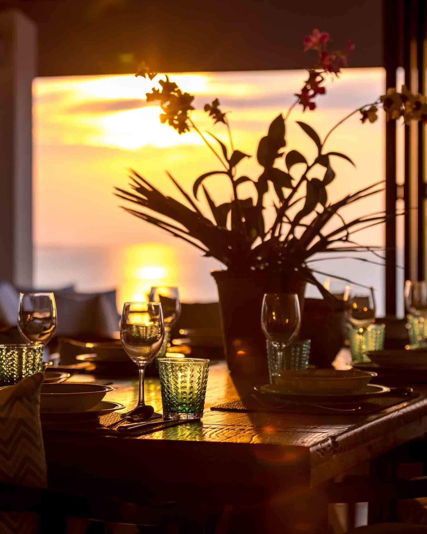 Intimate setting of a dining table beautifully arranged with wine glasses and floral centerpieces against a stunning sunset backdrop