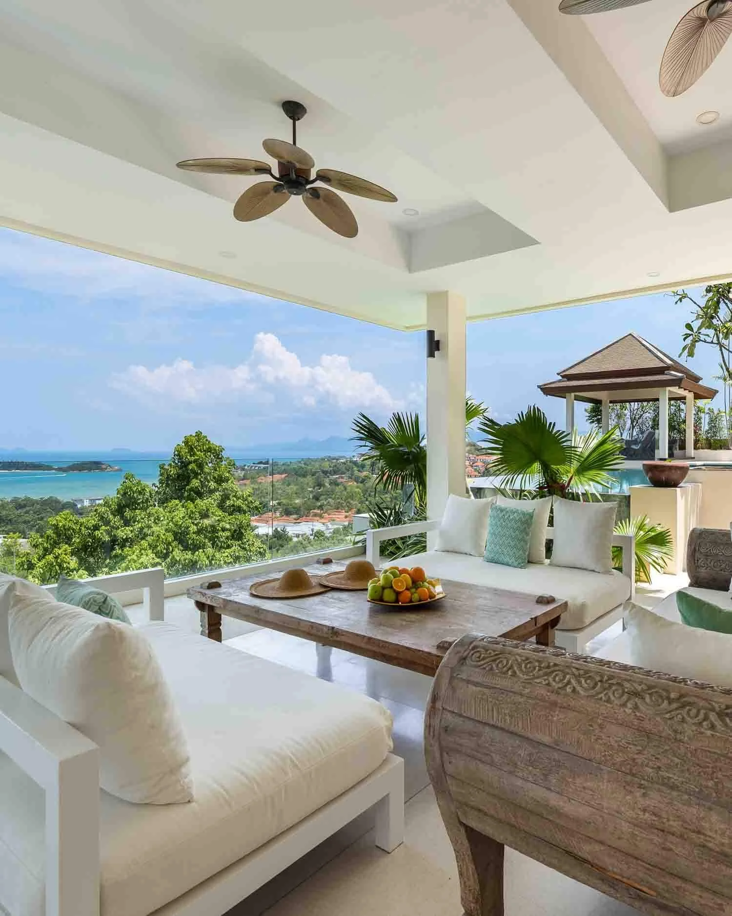 Shaded outdoor living lounge with white sofas and a rustic wooden coffee table framing views of the pool deck and sea