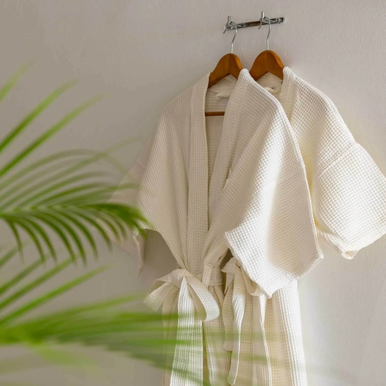 White waffle weave bathrobe with a belt hanging on a wall next to a green plant.