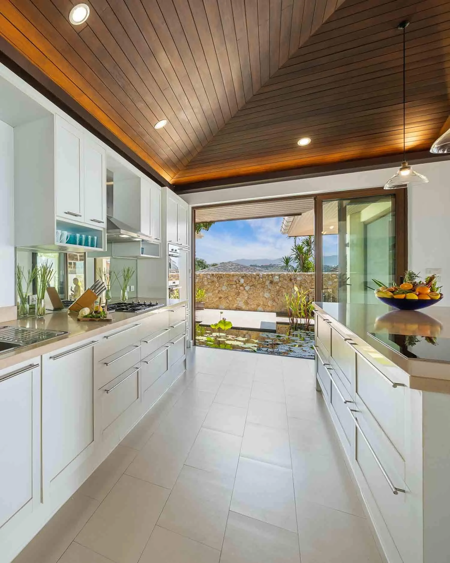 Modern white kitchen with sleek cabinetry and a breakfast bar, opening directly onto a tranquil outdoor stone garden water feature