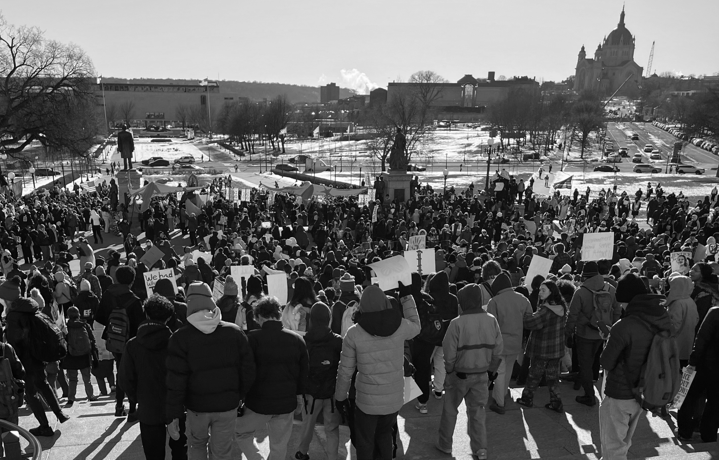Crowd of protesters holding signs and banners, gathered outdoors in a city square during daytime, with buildings and a large church or cathedral in the background.