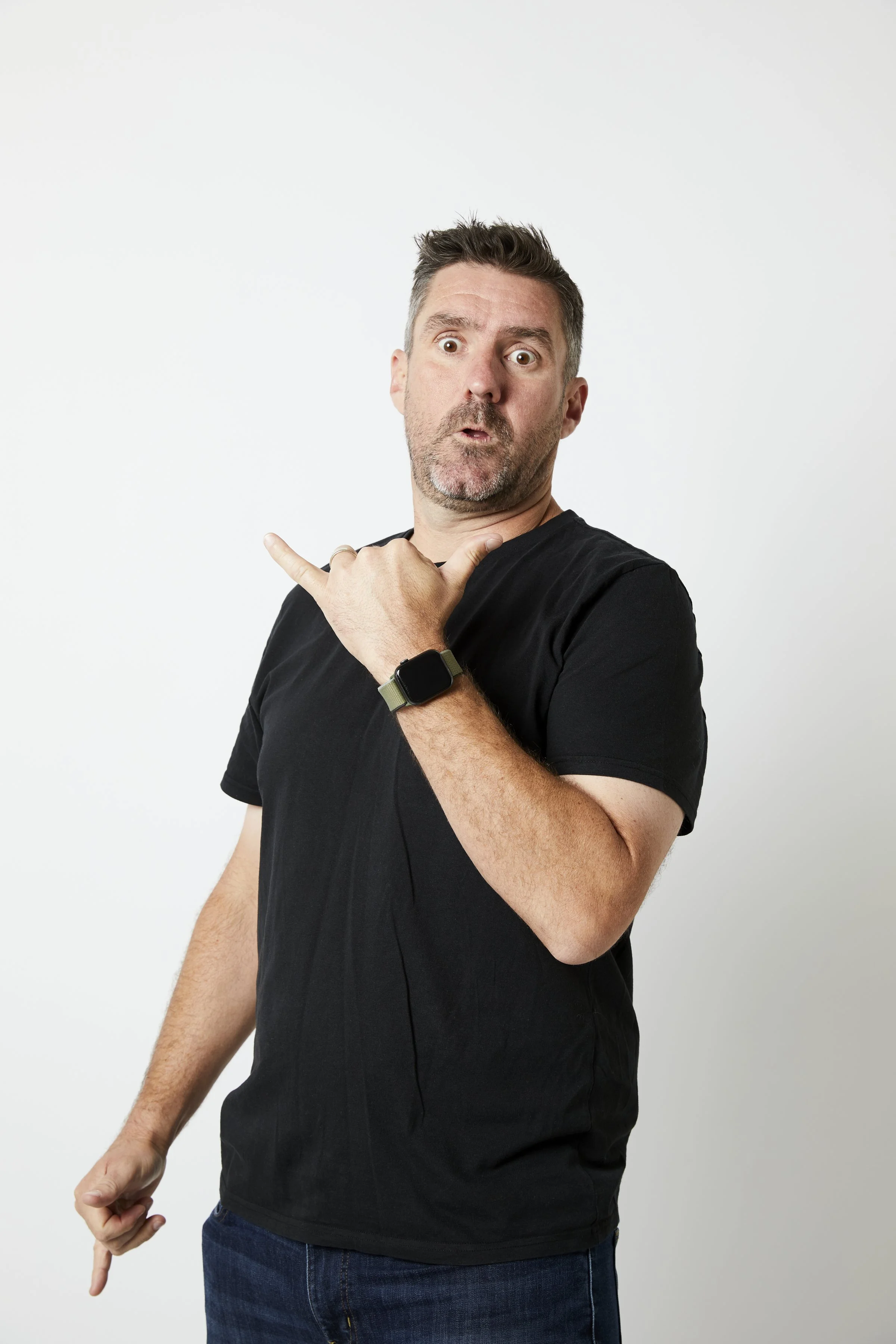 A man with short brown hair and a beard making a surprised face, pointing with his right hand to his left, wearing a black T-shirt and a smartwatch on his left wrist, standing against a plain white background.
