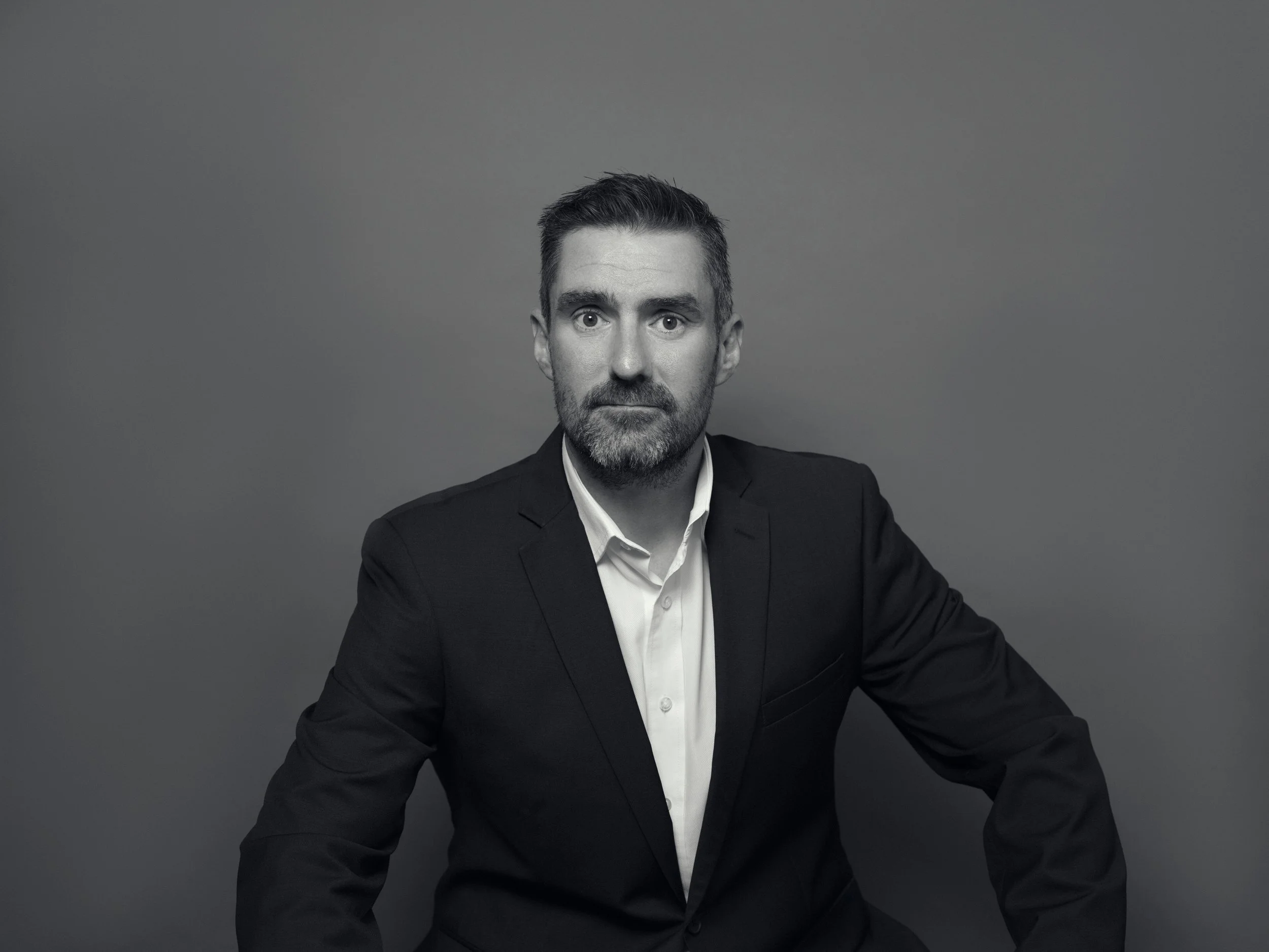 A black-and-white portrait of a man with short dark hair, a beard, wearing a dark suit and white shirt, looking directly at the camera against a plain gray background.
