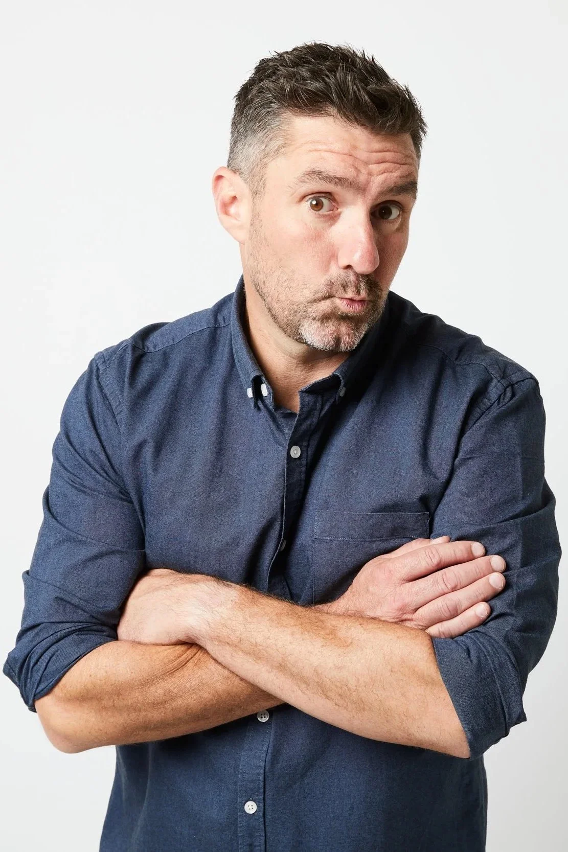 A man with short dark hair, a beard, and mustache, wearing a dark blue button-up shirt, standing with arms crossed, looking at the camera with a surprised expression against a plain white background.