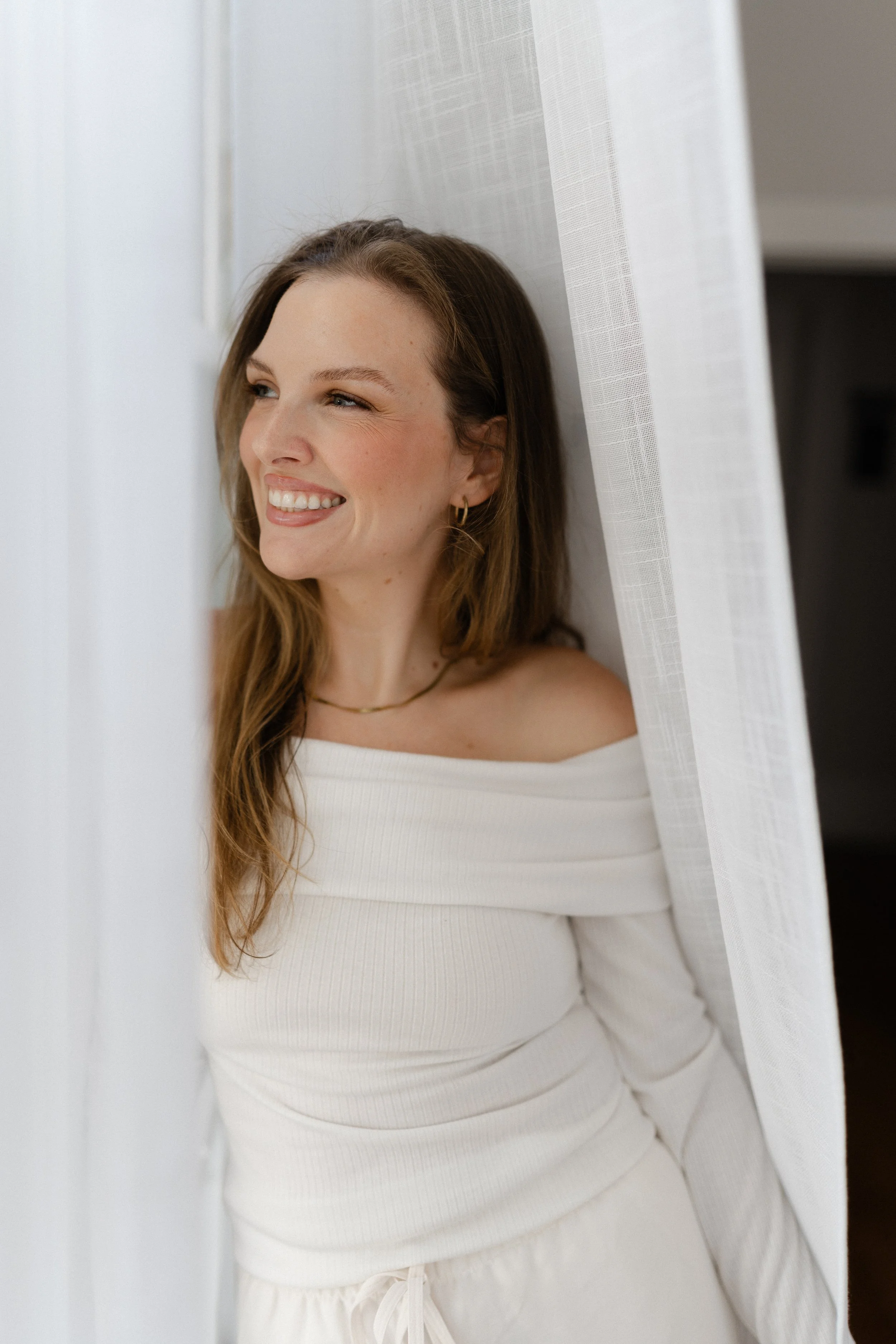 Smiling woman in a white dress standing behind sheer curtains