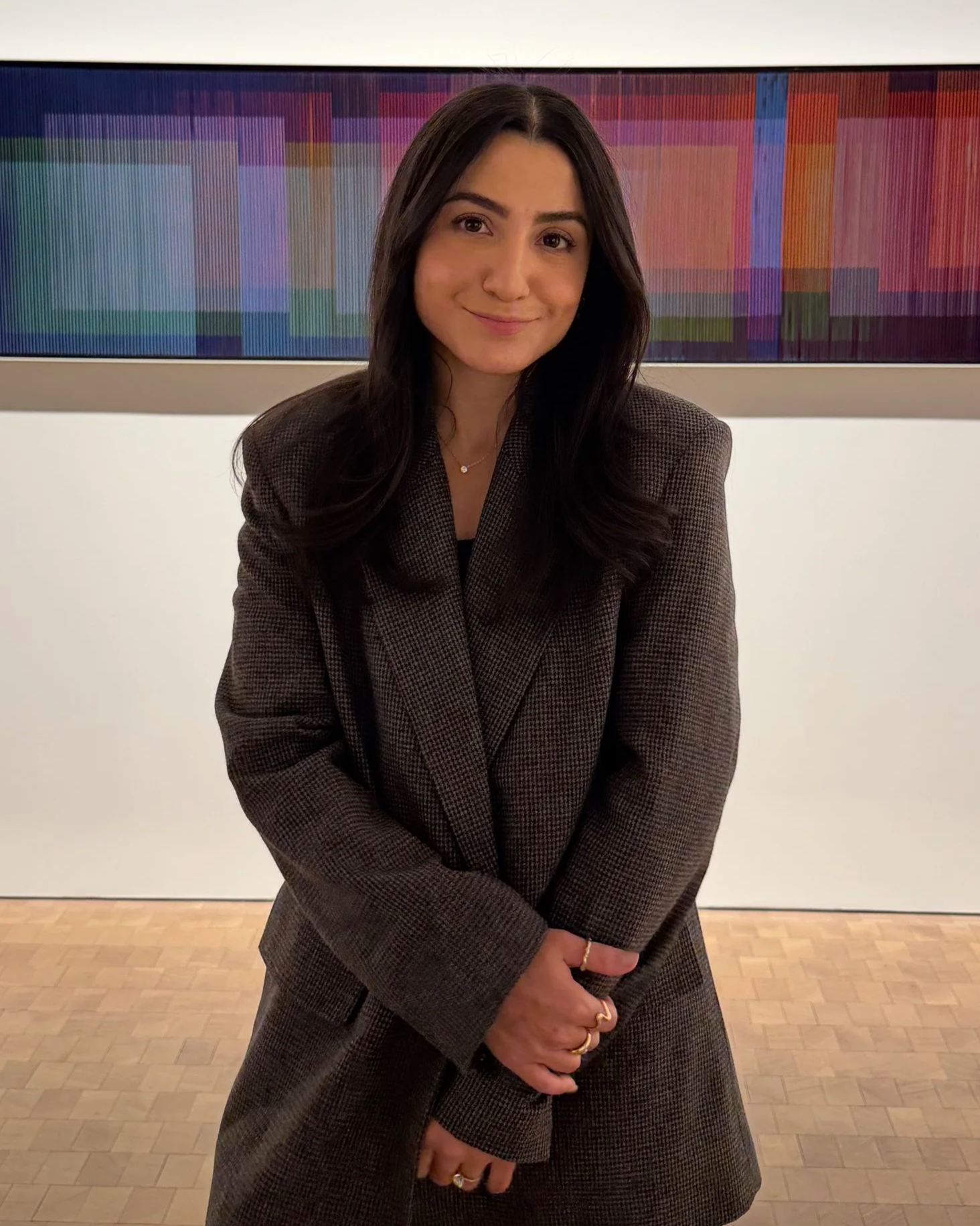 A woman with long dark hair and a checkered blazer standing in front of a colorful horizontal art piece, smiling softly with her hands clasped in front of her.