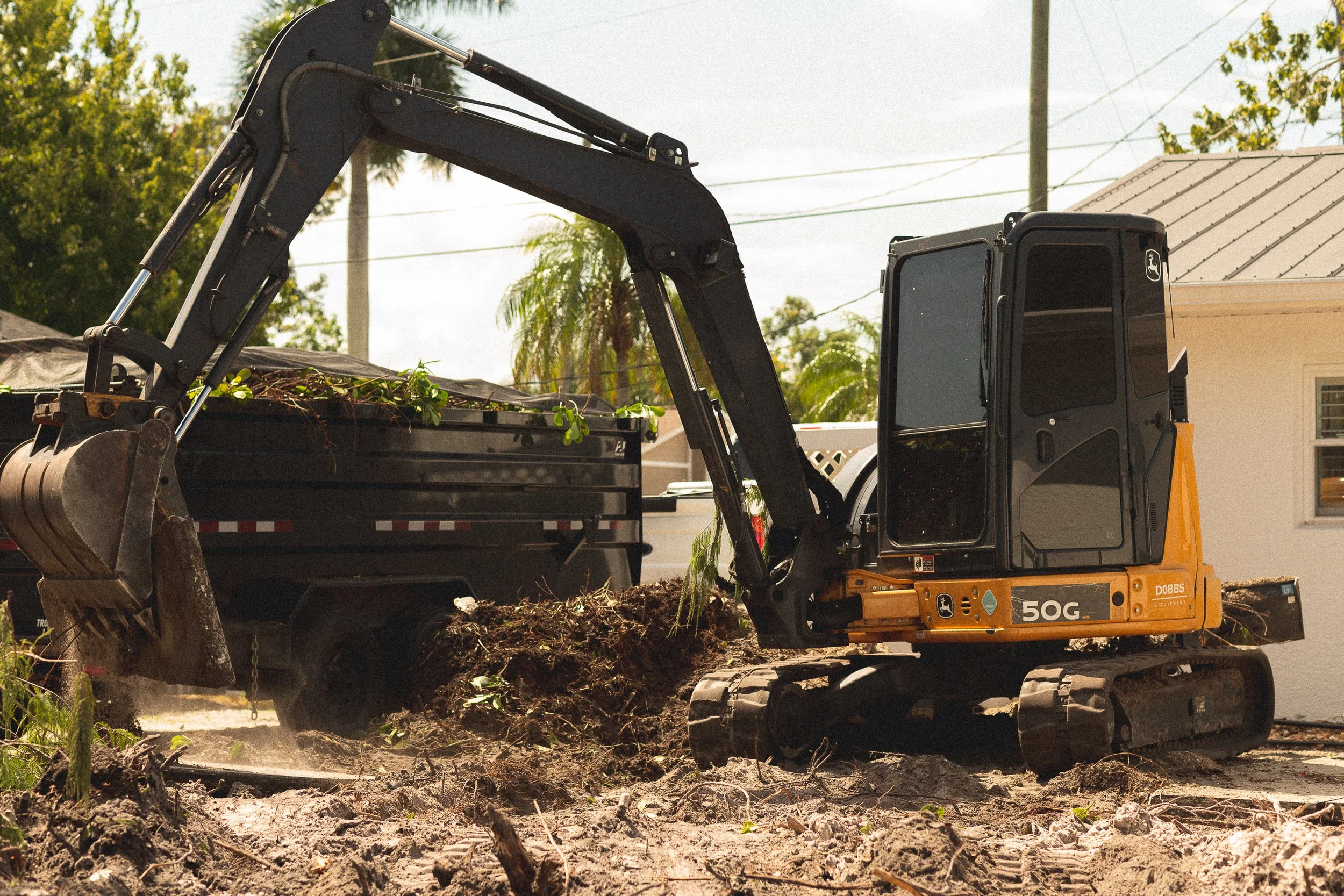 A yellow mini excavator digging into the ground next to a black truck filled with dirt and green plants, with trees and power lines in the background.