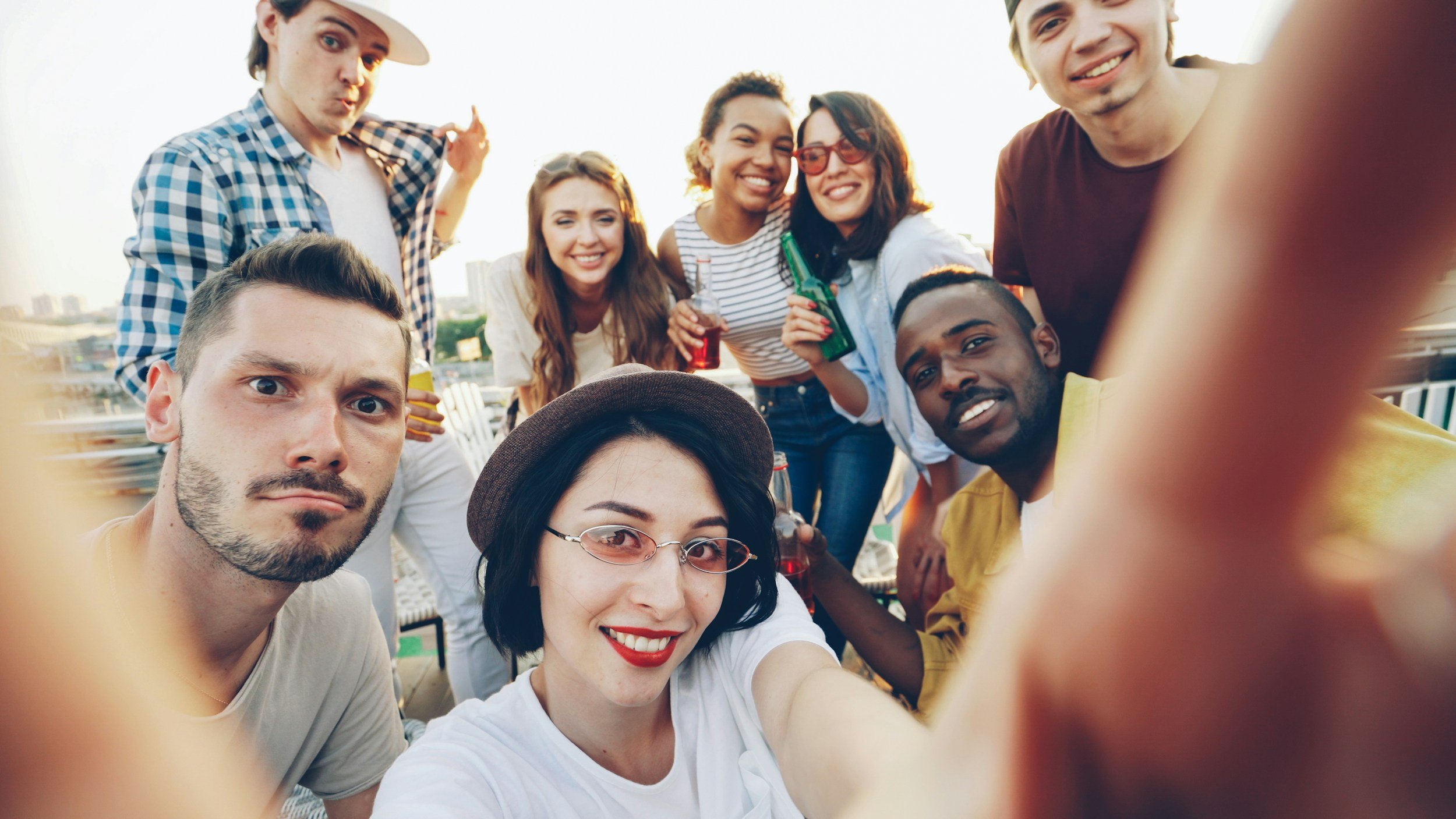 Group of young friends taking a selfie together at sunset, smiling and holding drinks, with buildings in the background.