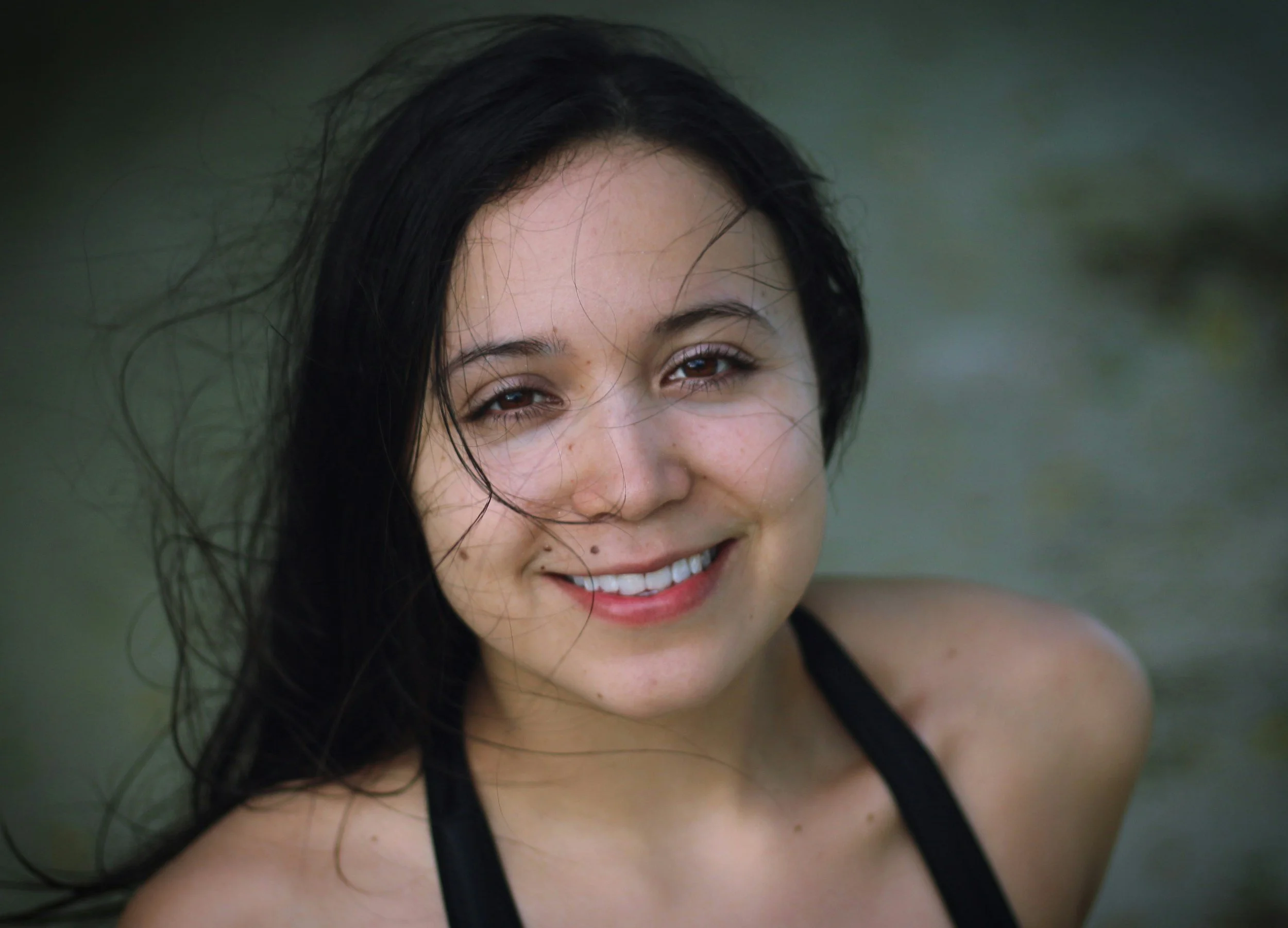 Close-up of a smiling woman with dark hair and brown eyes, wearing a black sports top, outdoors with a blurred green background.