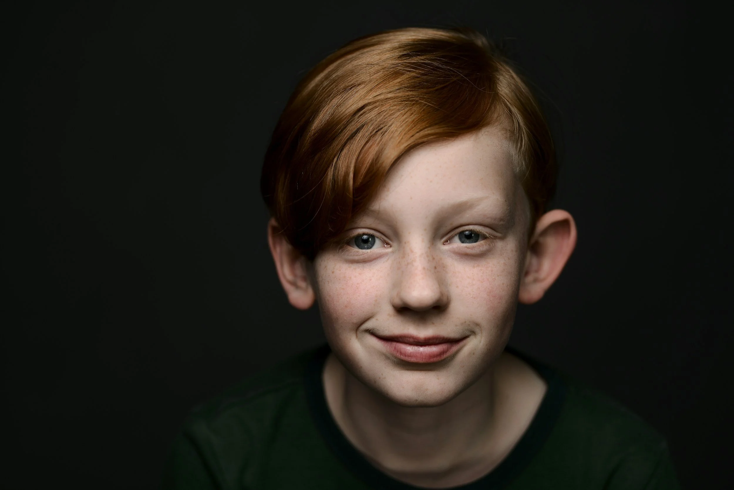 Close-up of boy with red hair, blue eyes, freckles, smiling against black background.