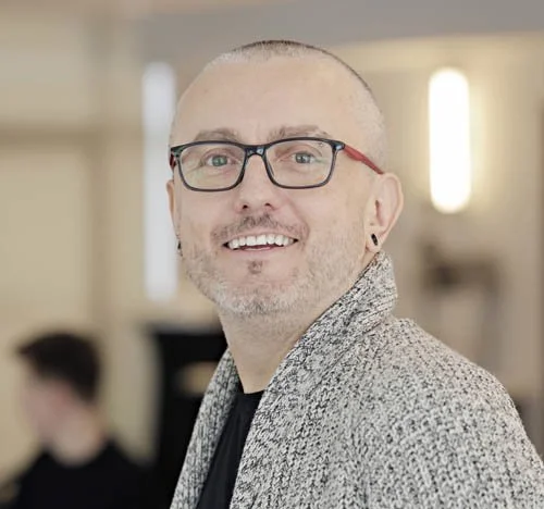 A smiling man with glasses and a shaved head, wearing a patterned blazer in an indoor setting.