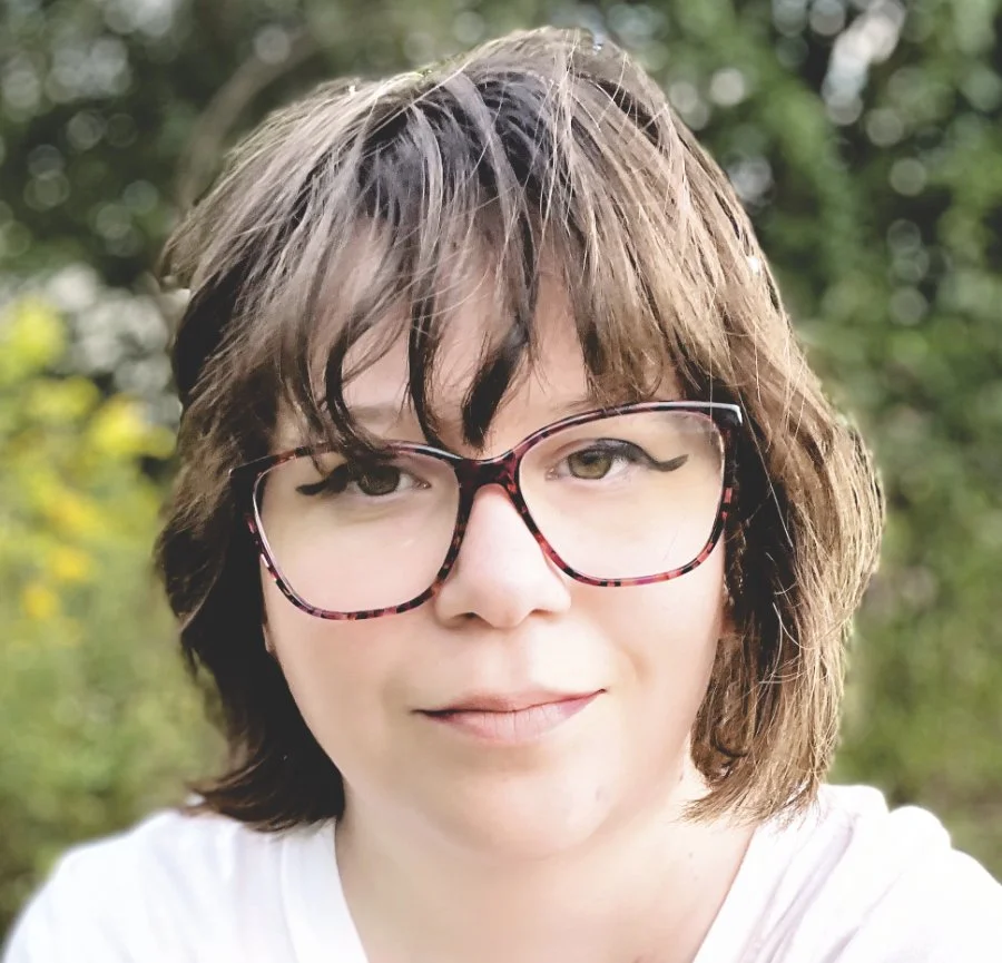 Close-up of a young person with brown, slightly messy hair, wearing pink and black glasses, outdoors with greenery blurred in the background.
