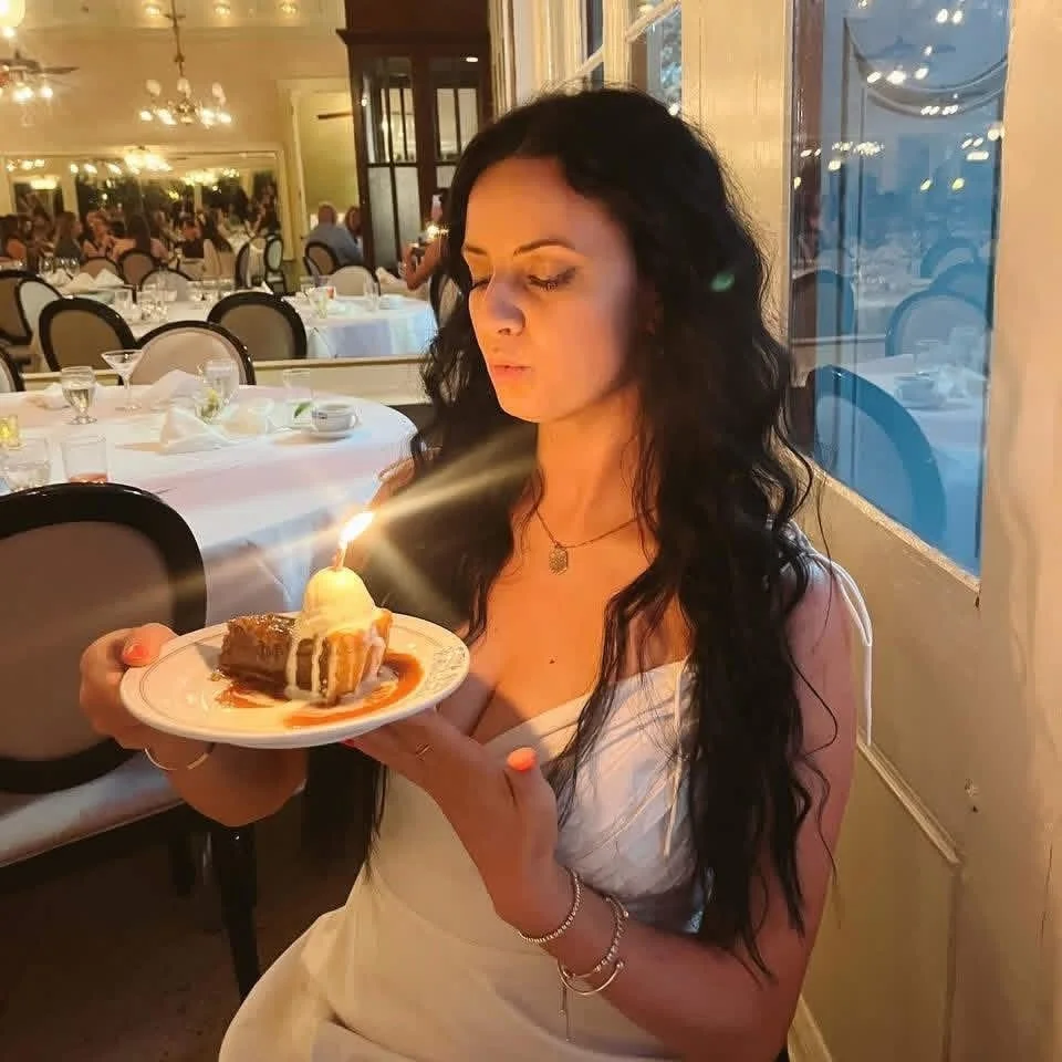 A woman with long dark curly hair and wearing a white dress is holding a plate with a slice of cake topped with a lit birthday candle.