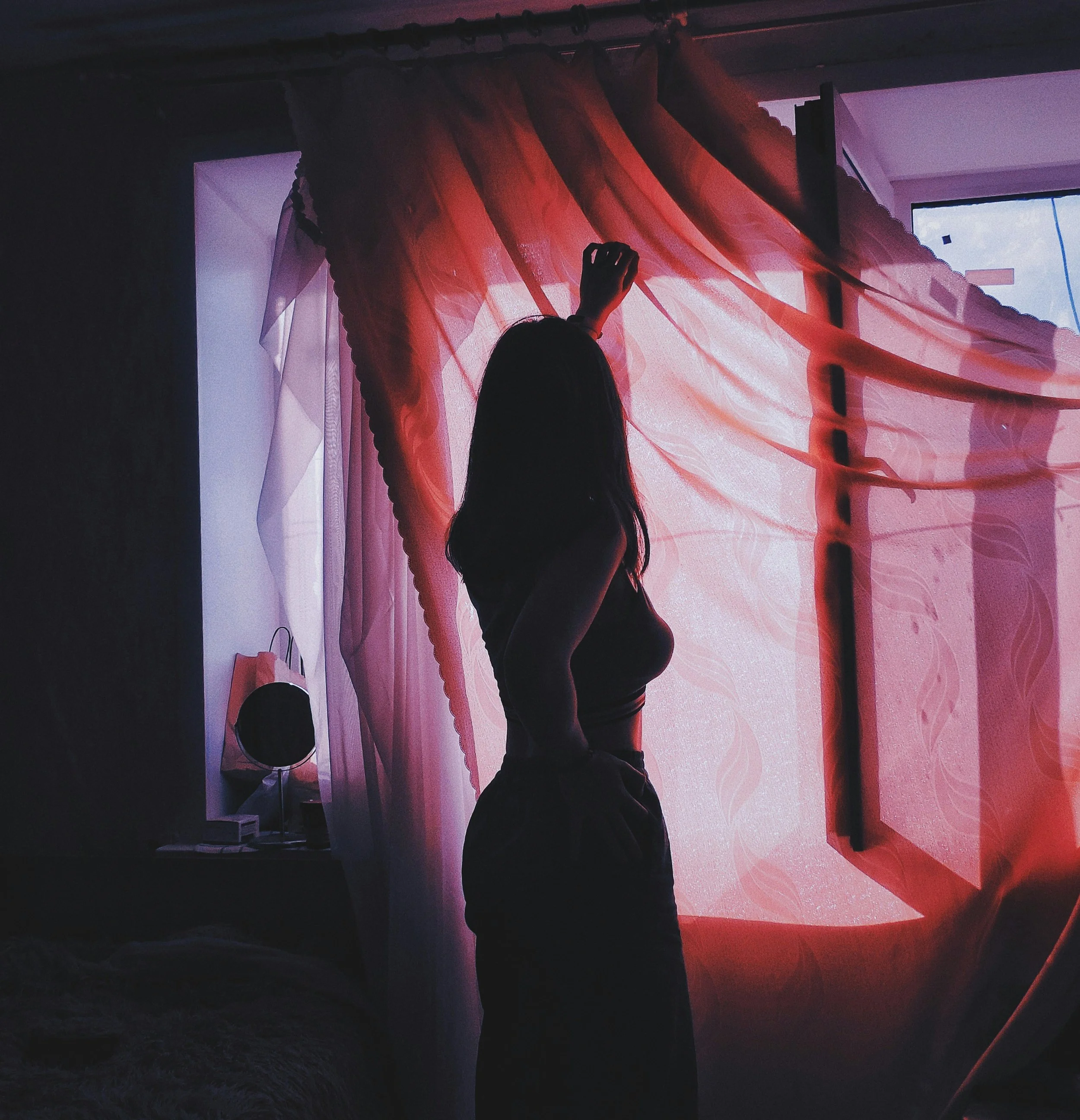 Silhouette of a woman with long hair adjusting red sheer curtains in front of a window with blue sky visible outside.