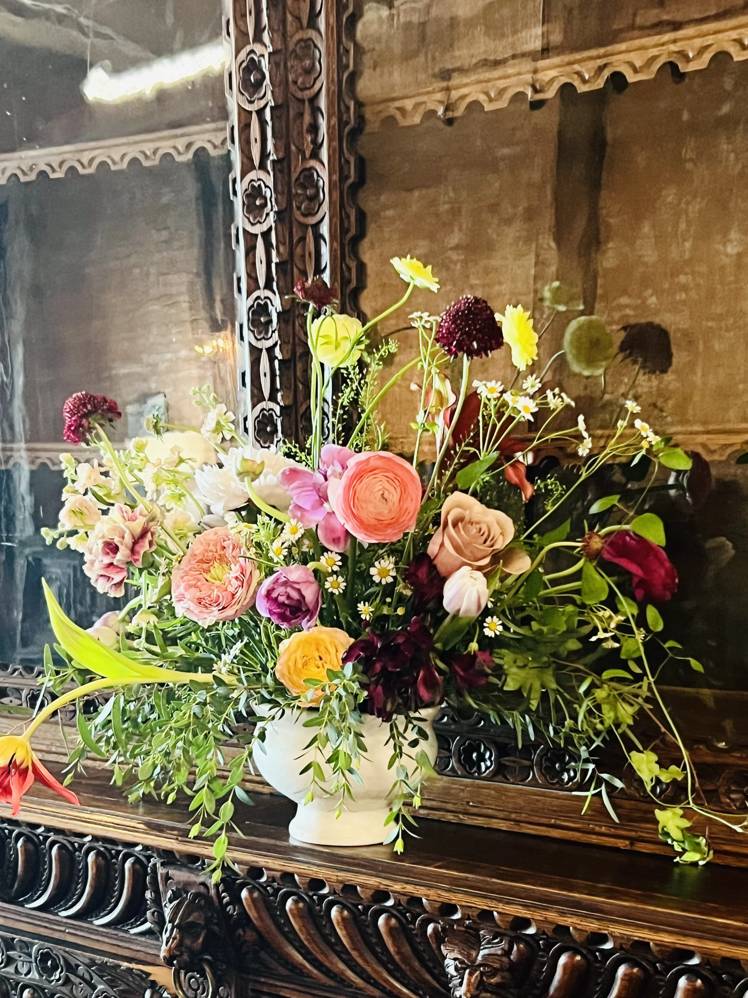 A colorful flower arrangement in a white ceramic vase sitting on a wooden table with an ornate carved dark wood frame behind it. The bouquet includes roses, ranunculus, daisies, and various greenery, against a rustic brick wall background.