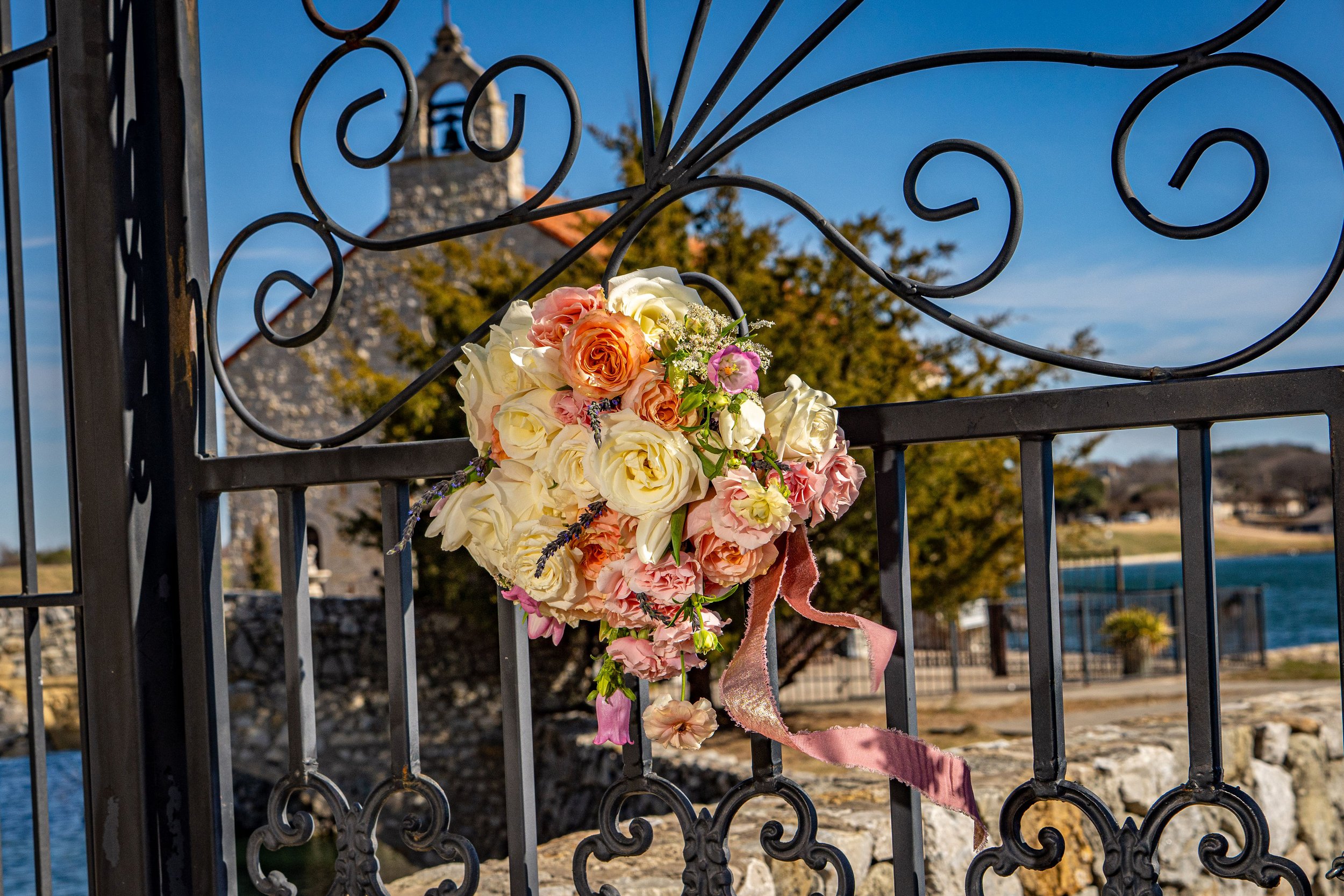 A floral arrangement attached to a black wrought iron gate, with a historic stone building and a body of water in the background on a clear day.