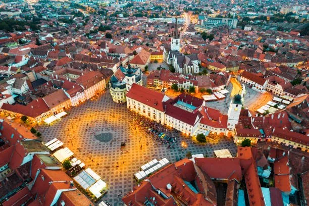 Aerial view of a European city square with historic buildings, a church, and a red-tiled roof, illuminated by streetlights at dusk.