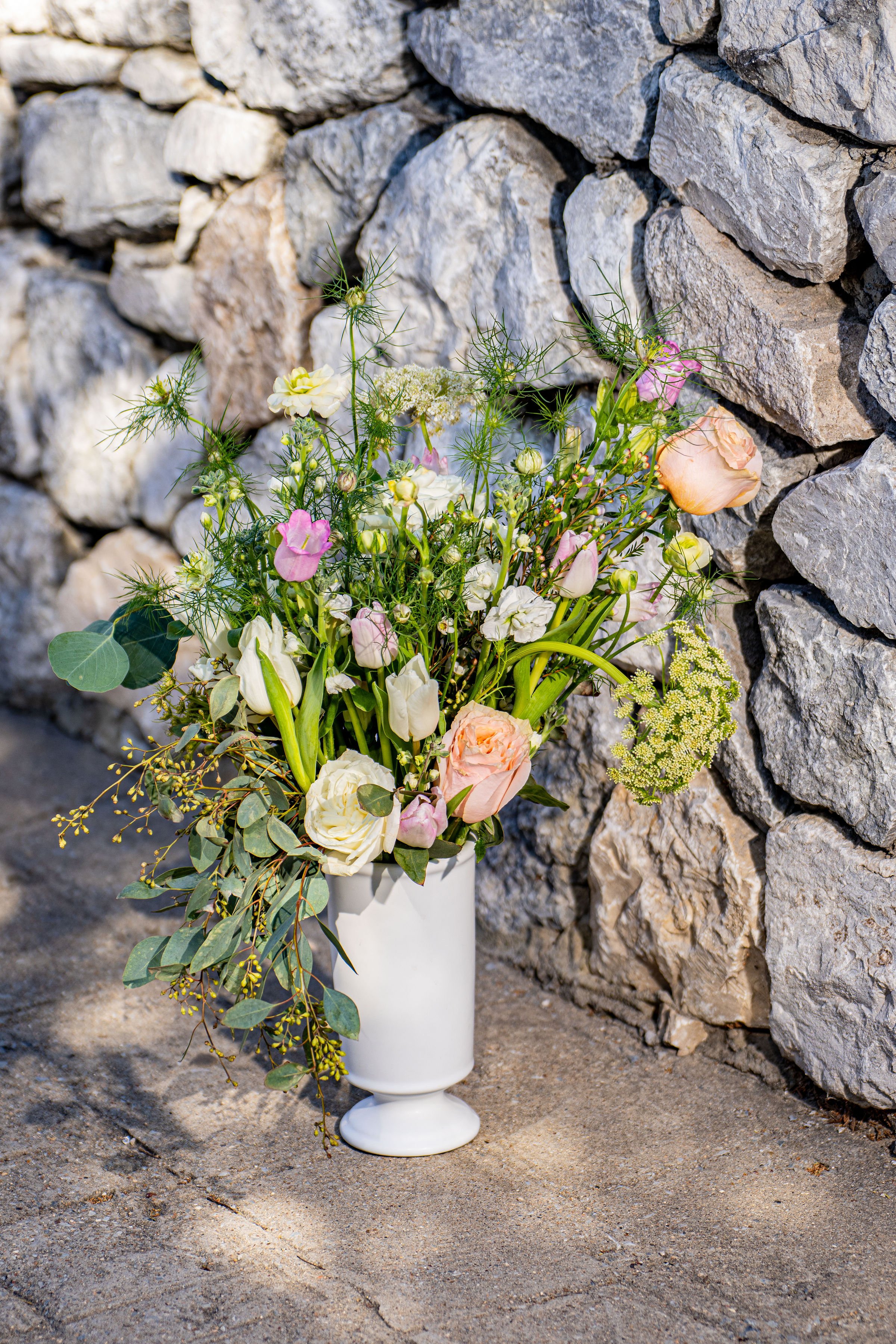 A bouquet of mixed flowers in a white vase placed on sandy ground with a stone wall background.