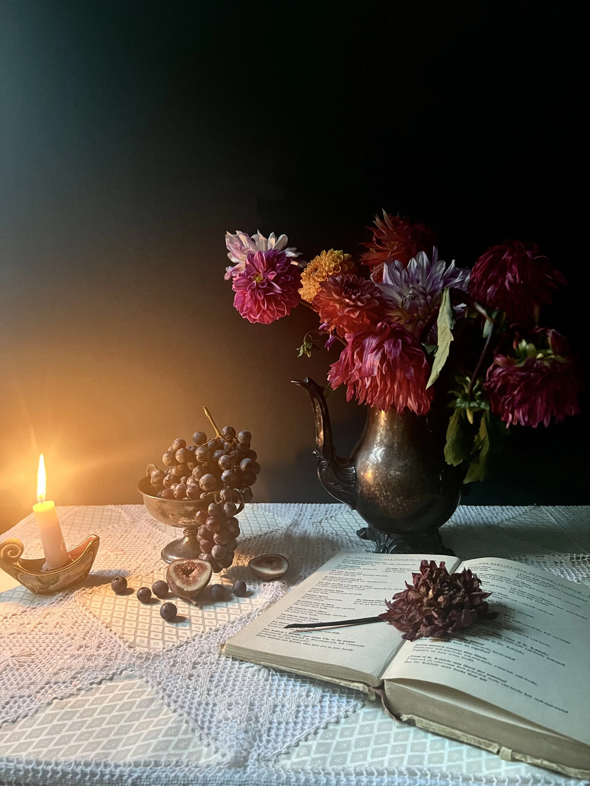 A still life of a table with a lit candle, a bowl of grapes, a cut fig, an open book, and a vase with pink and purple flowers, all set on a white lace tablecloth against a dark background.