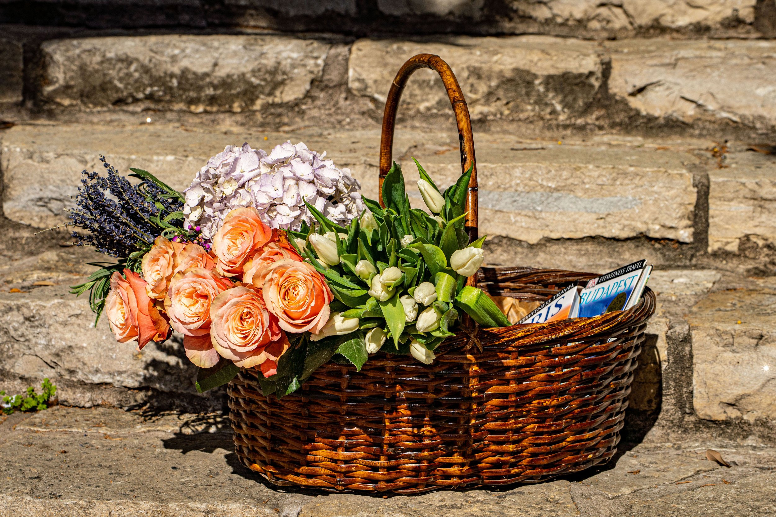 A wicker basket filled with white, peach, and purple flowers, including roses and tulips, resting on a stone surface with a stone wall background.