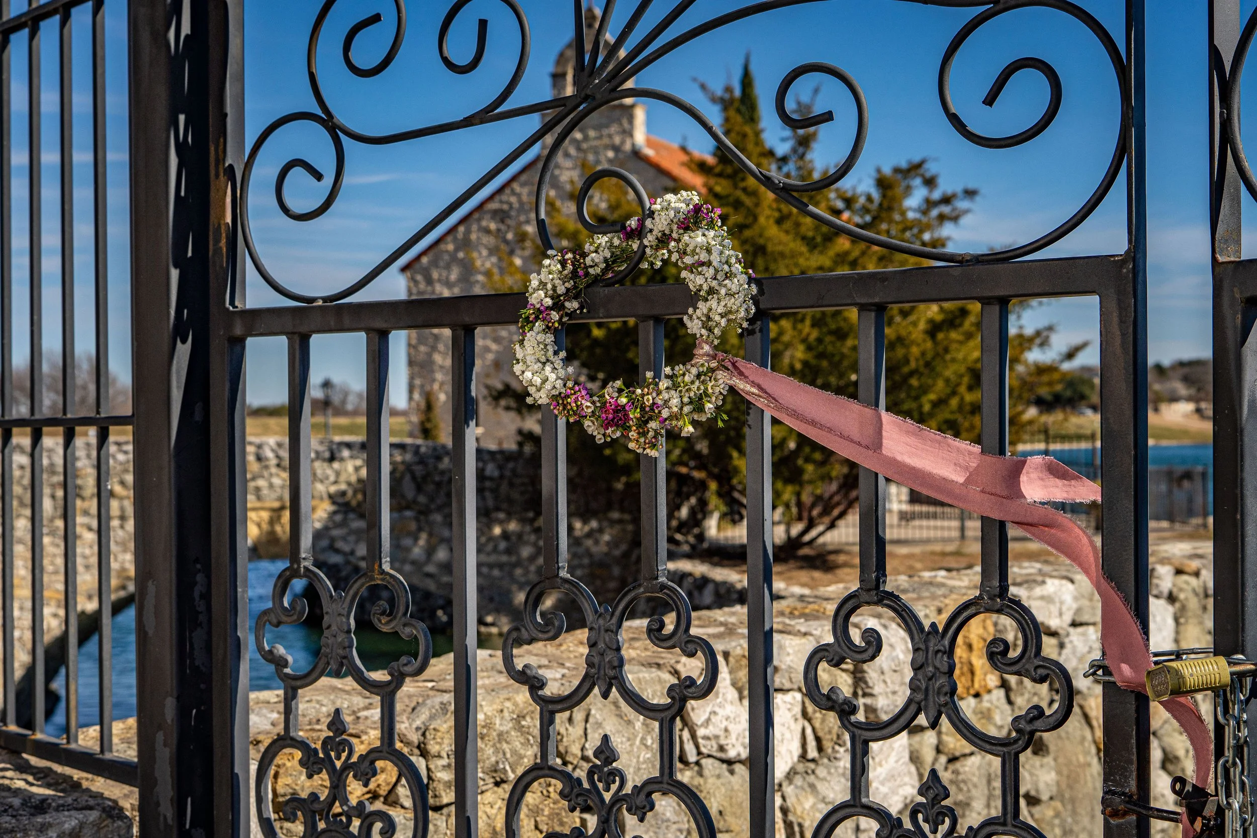 A decorative wrought iron gate with a floral wreath attached, a pink ribbon, and a padlock. In the background, there is a stone building with a reddish roof, trees, and a blue sky with some clouds.